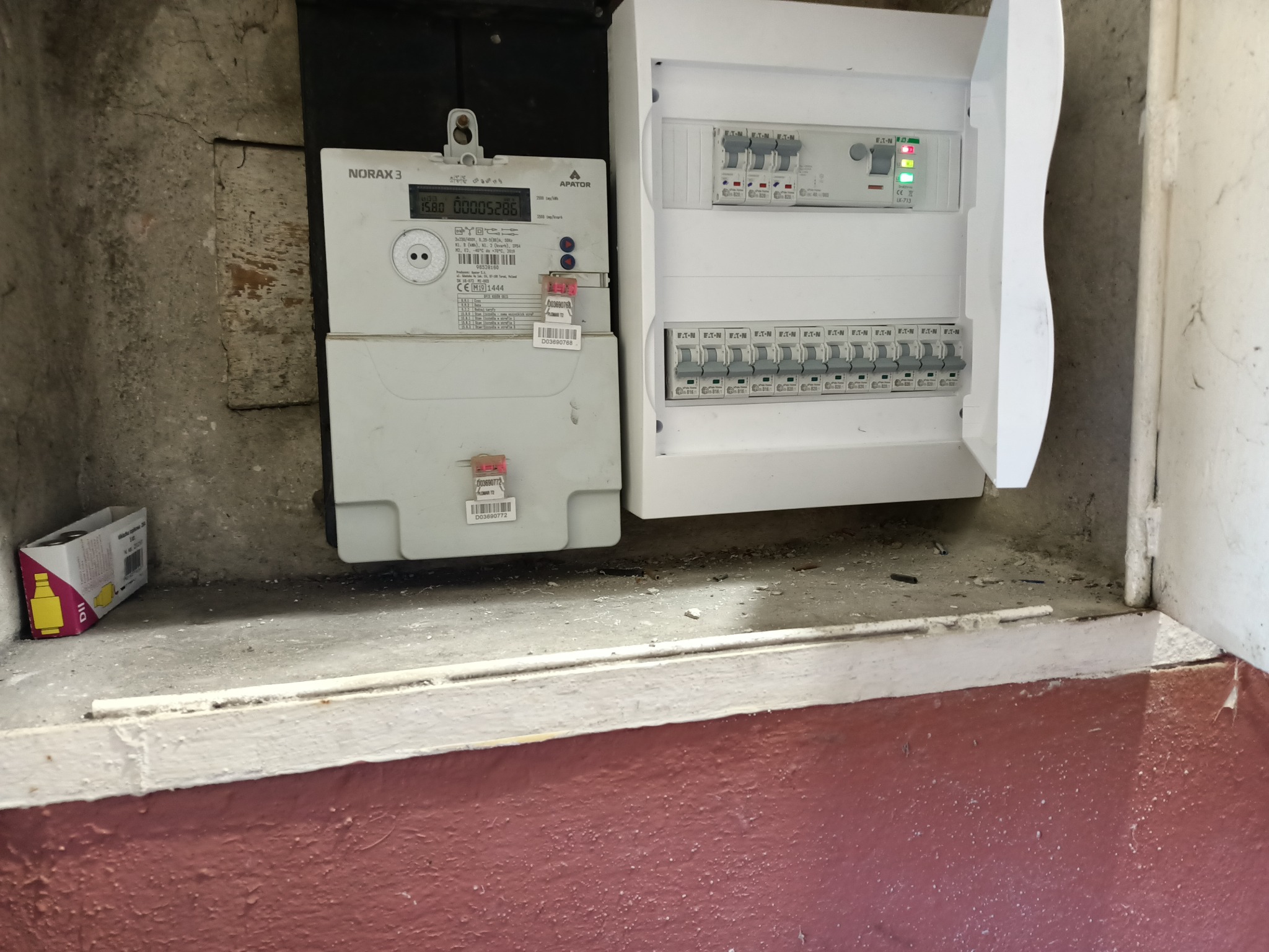 Licznik energii elektrycznej Norax 3 z wyświetlaczem LCD oraz biała skrzynka z bezpiecznikami, zamontowane na szarej, betonowej ścianie. Widoczna również kartonowa paczka obok licznika.