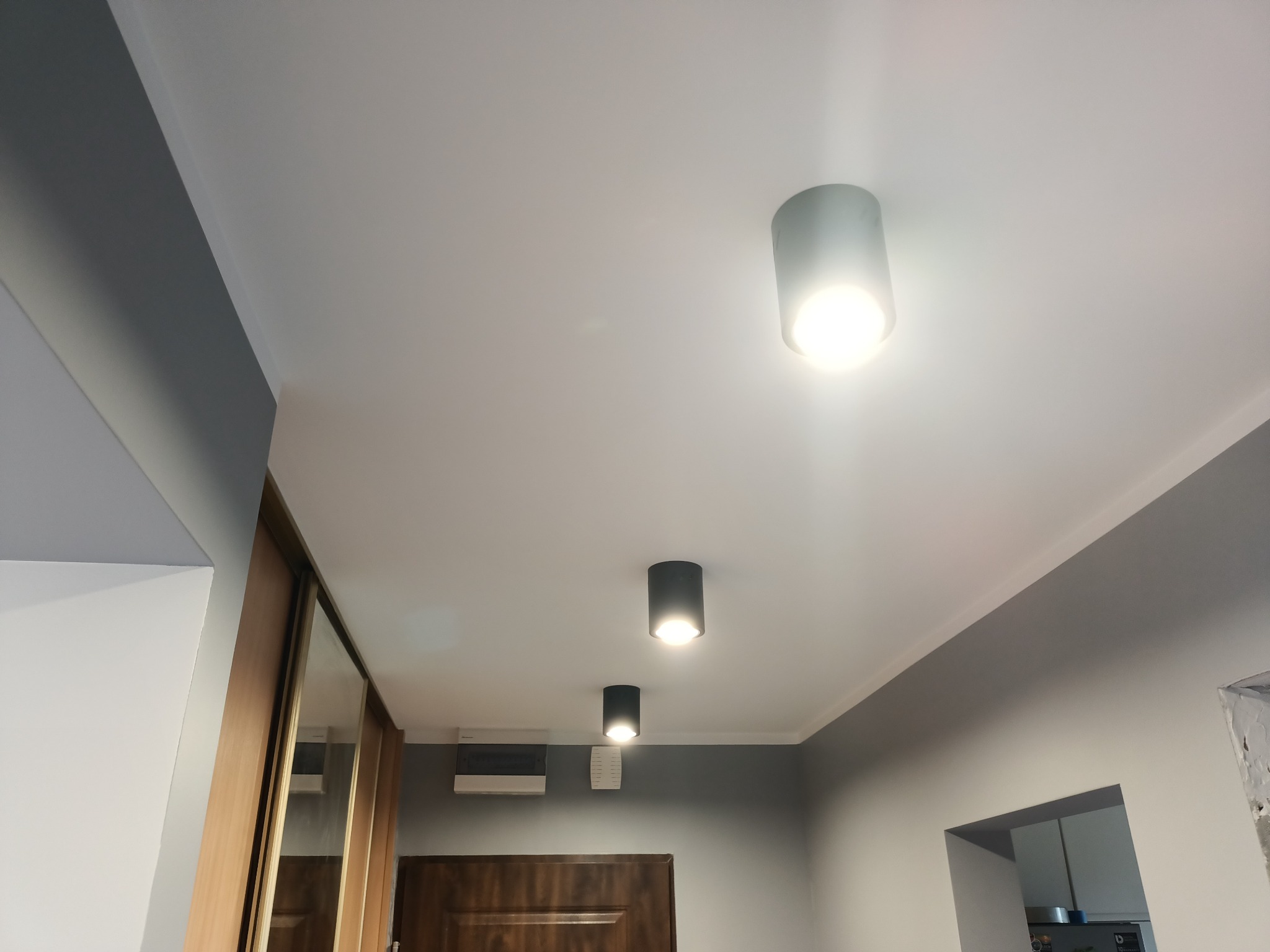 Trzy czarne lampy sufitowe tubowe LED, emitujące ciepłe światło, zamontowane na białym suficie w pomieszczeniu z szarą ścianą i drewnianymi elementami zabudowy.