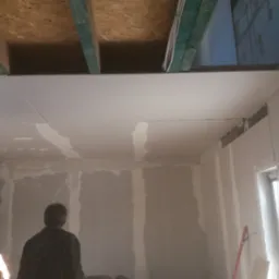 Montaż płyt kartonowo-gipsowych na suficie i ścianach w trakcie prac wykończeniowych, widoczne drewniane belki stropowe i fragment okna.