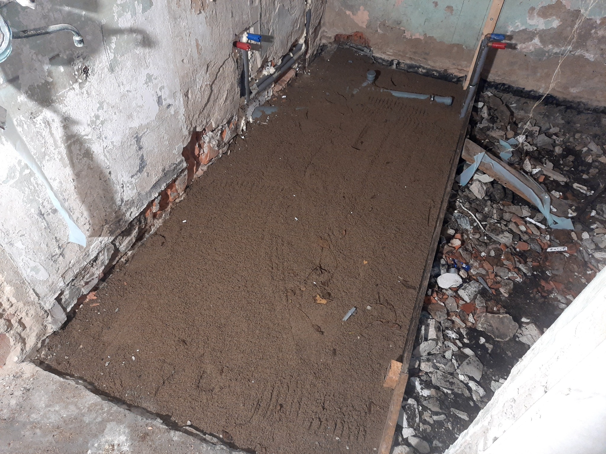 Przygotowanie podłoża pod wylewkę w pomieszczeniu: wyrównana warstwa piasku na podłodze z widocznymi instalacjami hydraulicznymi i fragmentami gruzu.