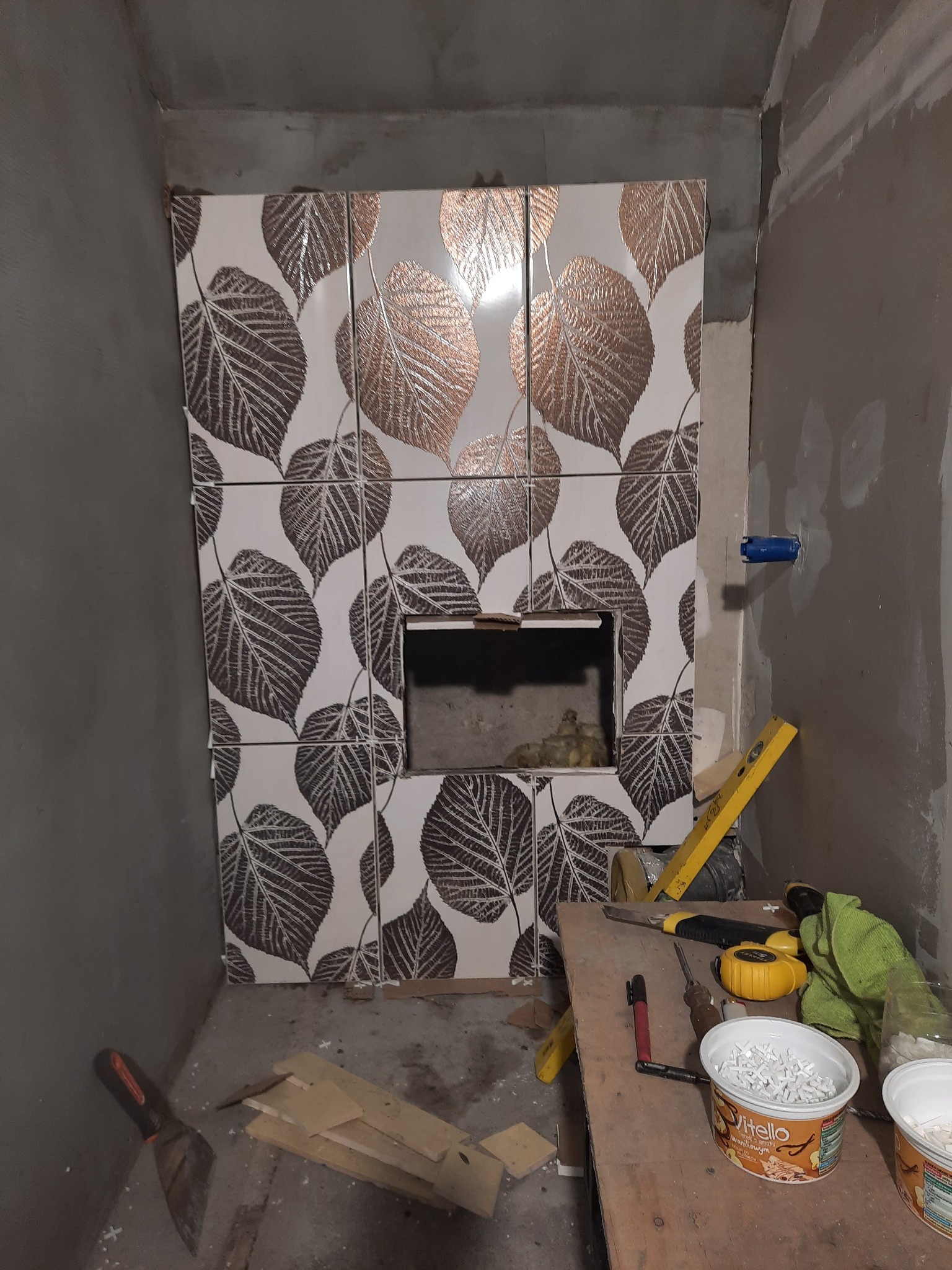 Fragment ściany w trakcie wykańczania z dekoracyjnymi płytkami w liście, widoczny otwór w ścianie, narzędzia glazurnicze na drewnianym stole oraz surowe ściany w tle.