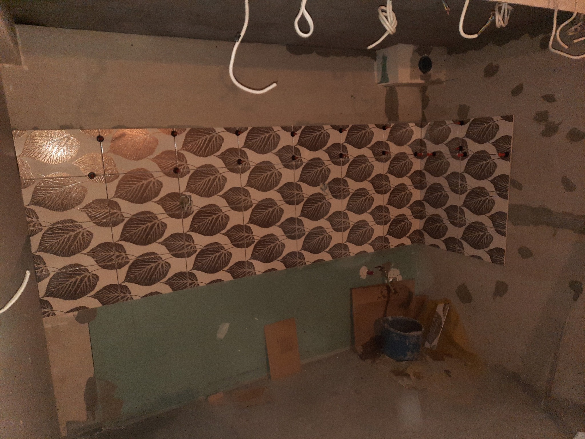 Ściana w trakcie wykańczania z ułożonymi płytkami w liście, widoczne przewody elektryczne, nieotynkowane ściany, wiadro i fragment płyty karton gips.