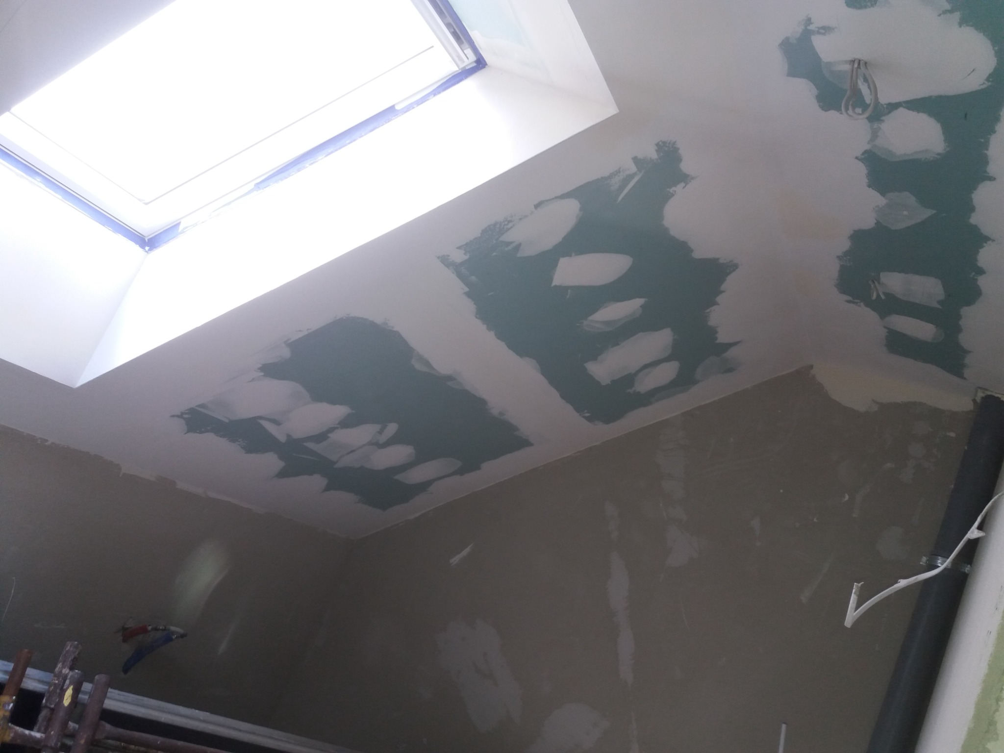 Sufit z oknem dachowym w trakcie szpachlowania, widoczne fragmenty nałożonej gładzi gipsowej, instalacja elektryczna wystająca ze ściany, fragment rusztowania w dolnej części kadru.
