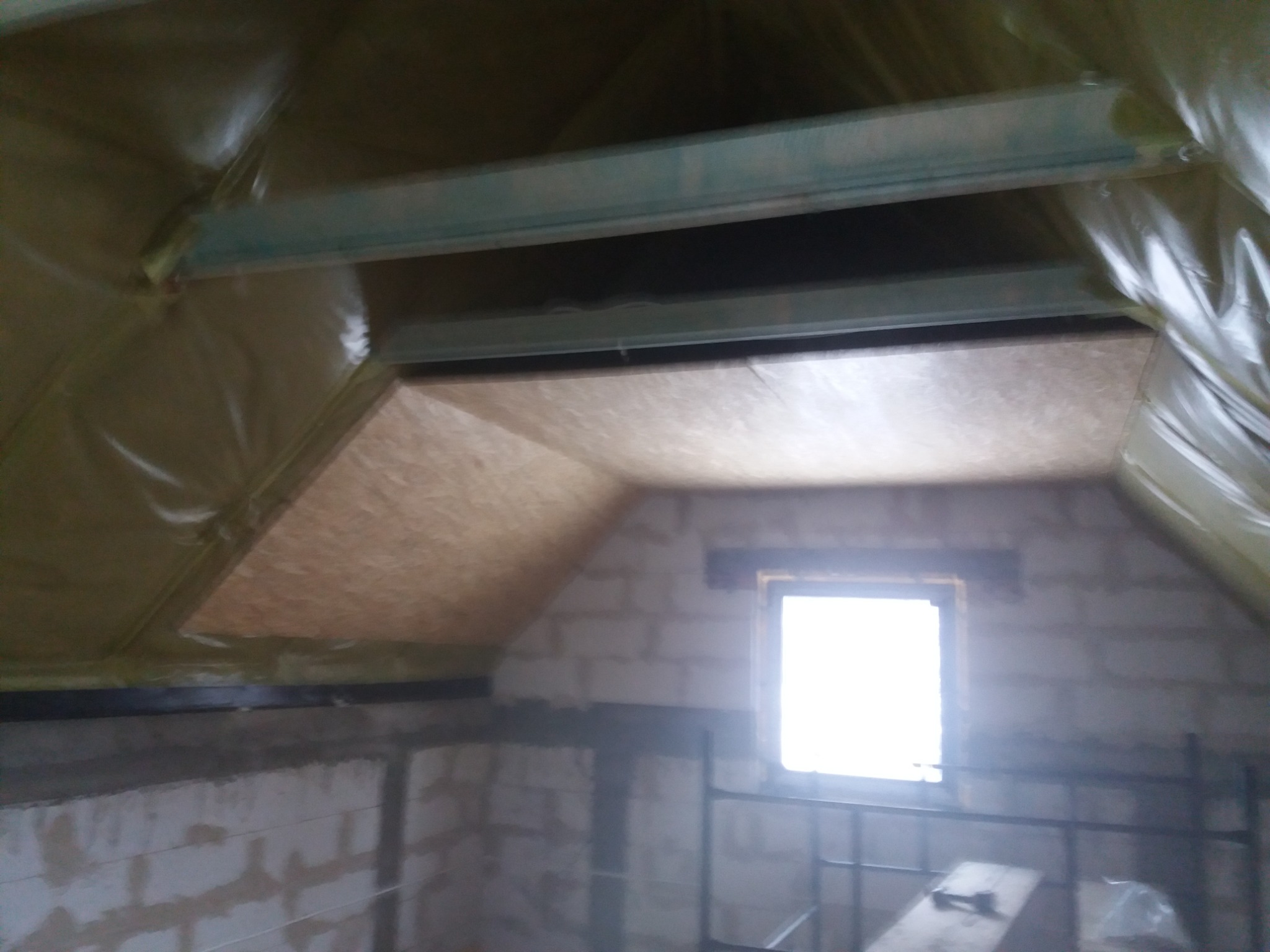 Poddasze w trakcie adaptacji: widoczne ocieplenie folią, konstrukcja metalowa, fragment płyty OSB na skosie dachu oraz okno w surowej ścianie z bloczków.