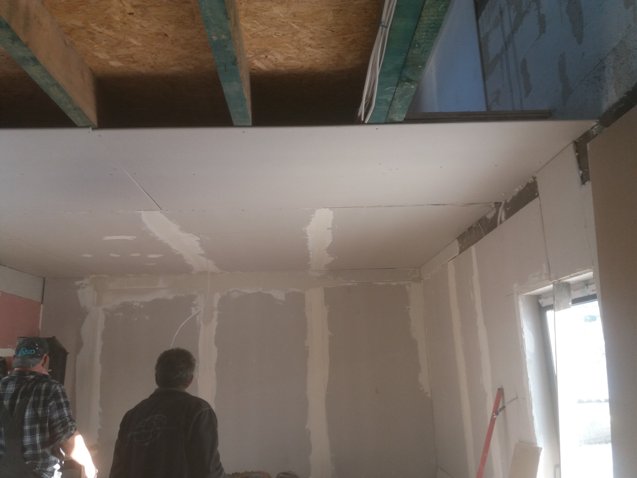 Montaż płyt kartonowo-gipsowych na suficie i ścianach w trakcie prac wykończeniowych, widoczne drewniane belki stropowe i fragment okna.