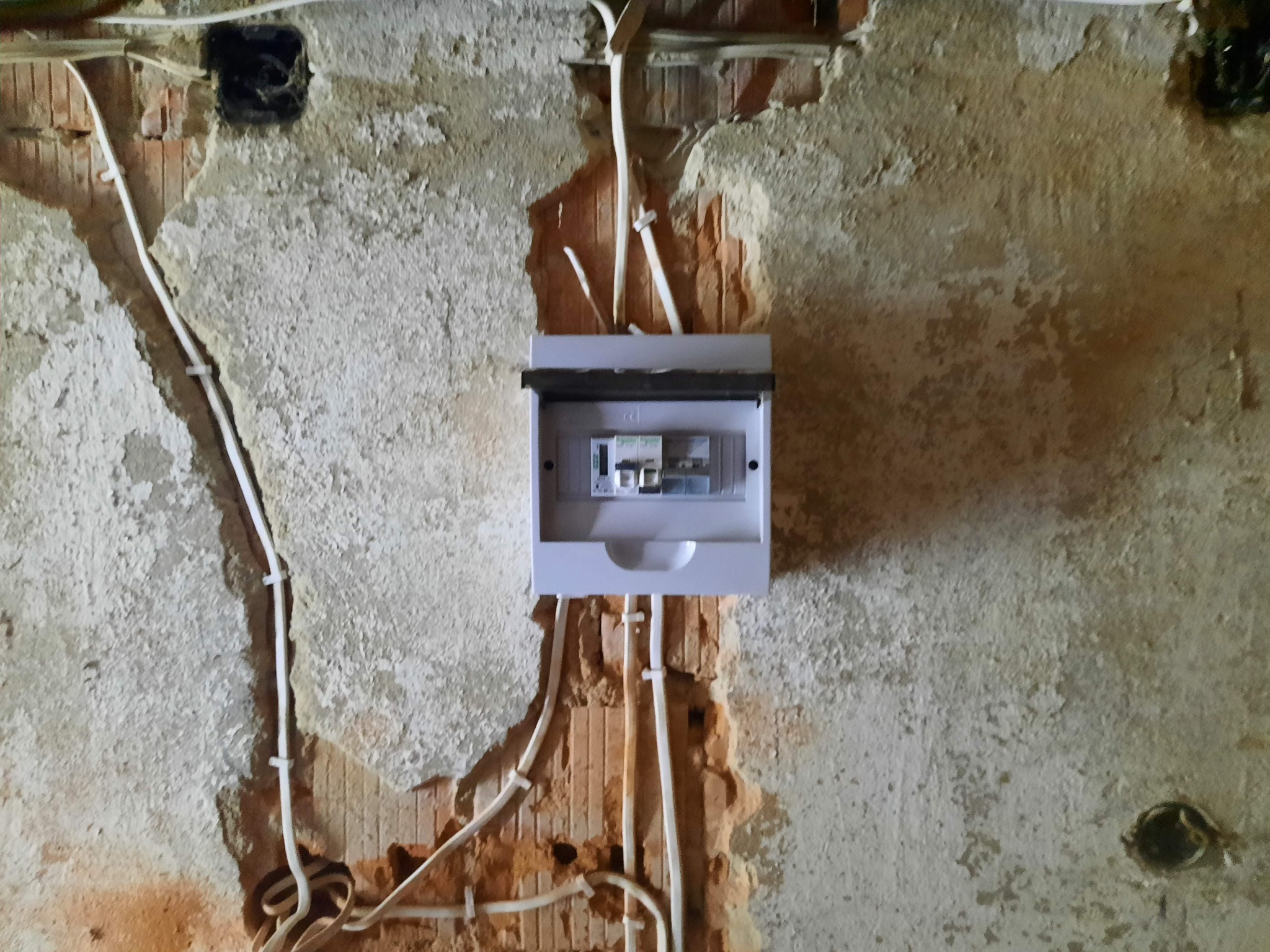 Montaż skrzynki elektrycznej z bezpiecznikami na starej, uszkodzonej ścianie z widoczną cegłą i przewodami elektrycznymi.