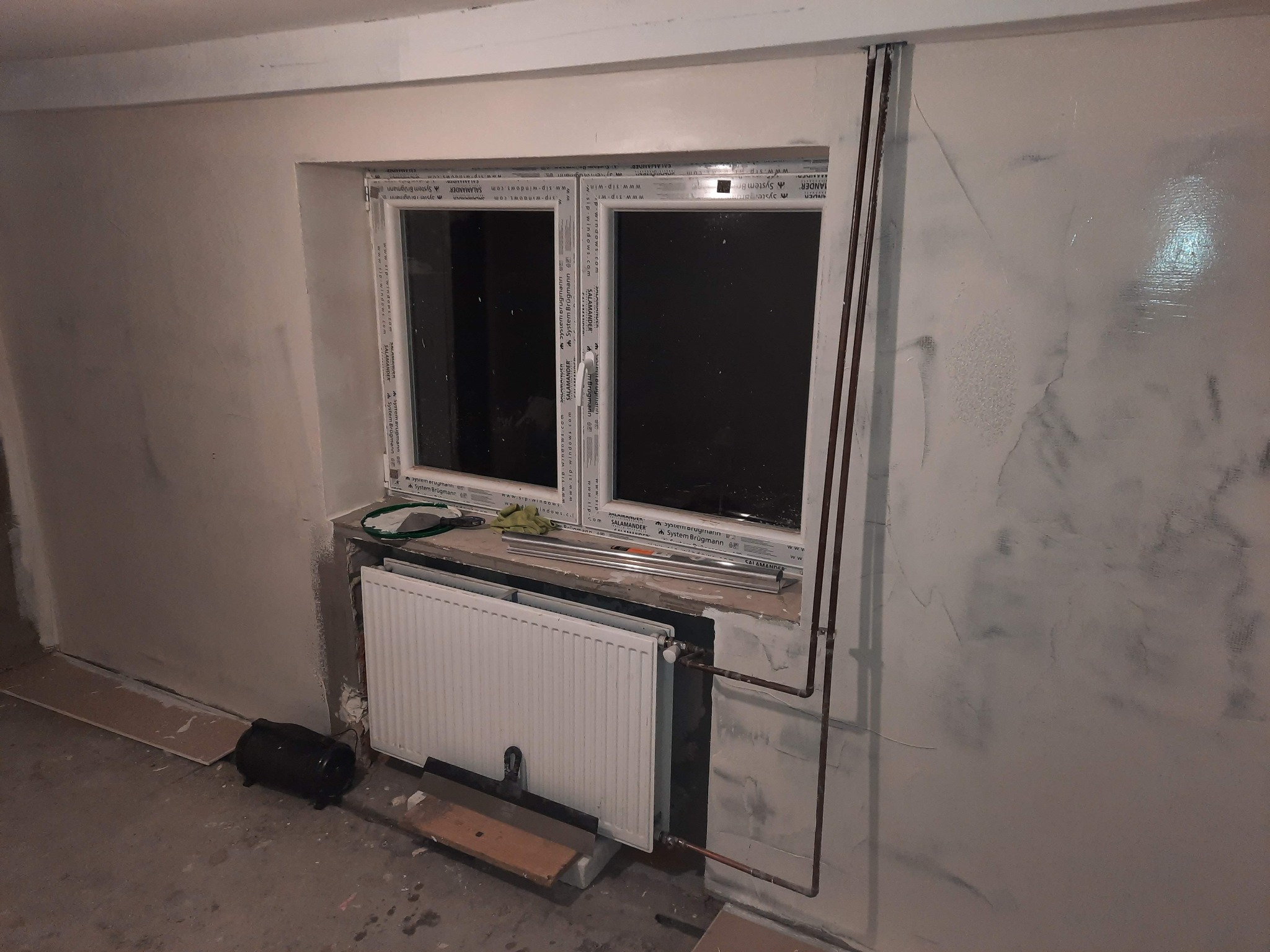 Świeżo pomalowane ściany z efektem dekoracyjnym, okno PCV z parapetem, grzejnik, rury grzewcze i narzędzia malarskie w surowym wnętrzu.