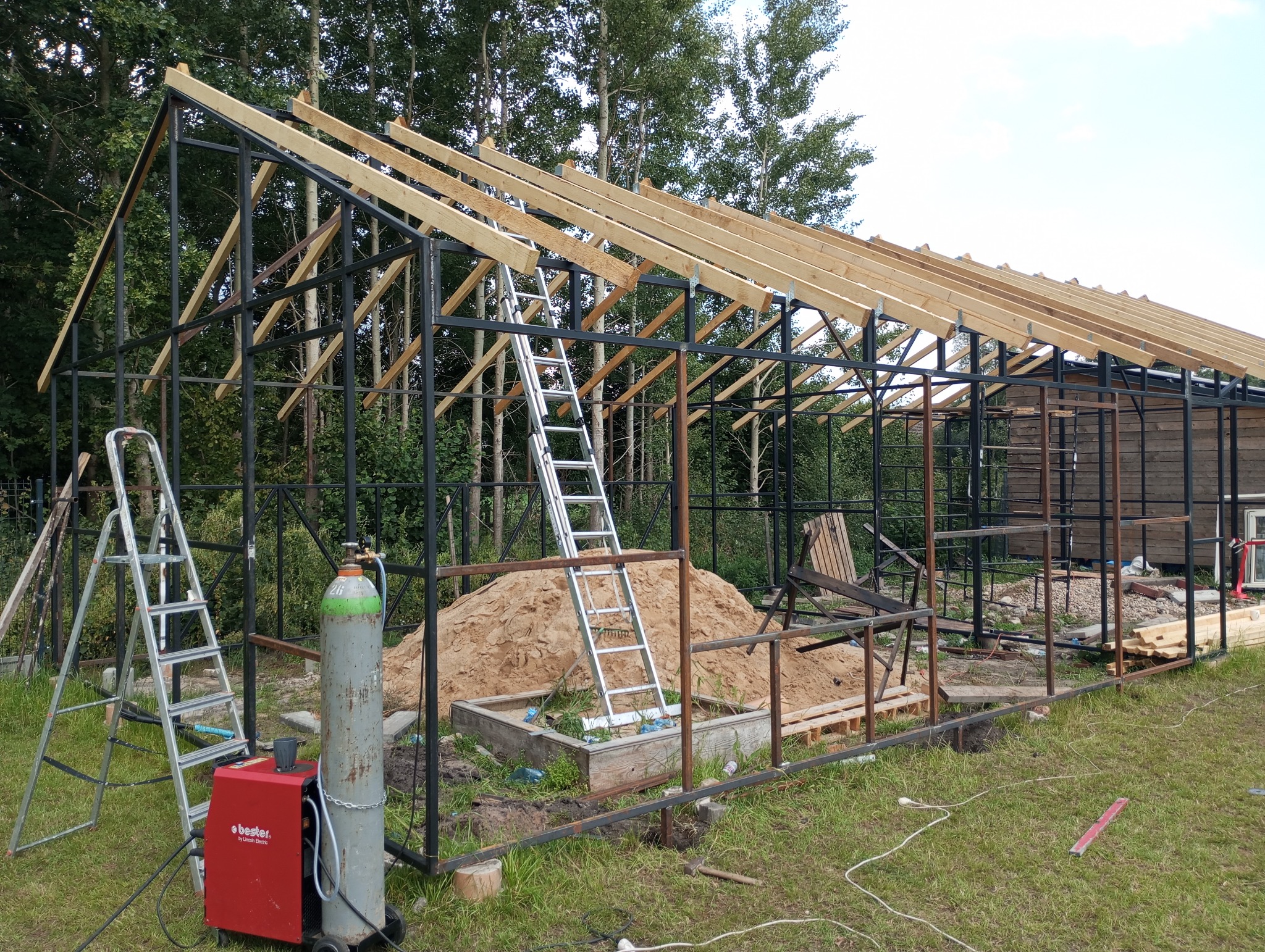 Konstrukcja szkieletowa budynku w trakcie budowy, widoczna spawarka, butla z gazem, drabiny i pryzma piasku.