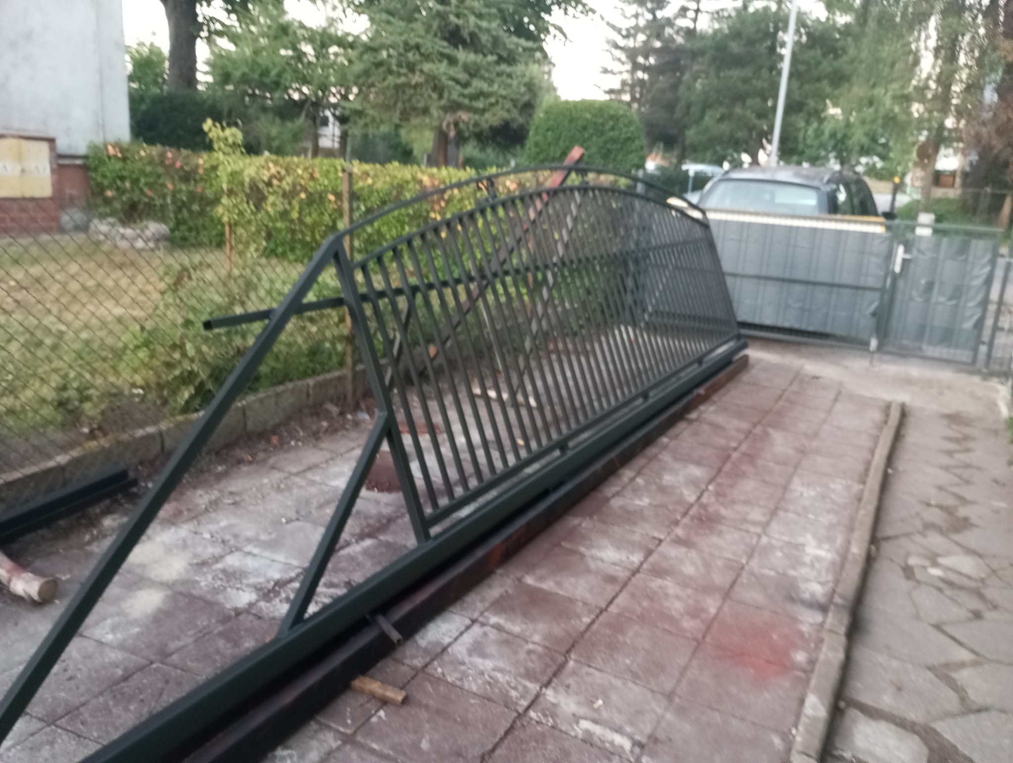 Niezamontowany segment bramy posesyjnej z metalowymi prętami i ramą, leżący na chodniku z kostki, w tle widoczny fragment ogrodzenia i samochód osobowy.