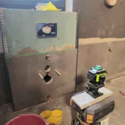 Montaż stelaża podtynkowego Geberit Omega z zielonym laserem krzyżowym ustawionym na wiadrze i pojemniku, widoczne otwory na instalację sanitarną i fragment ułożonych płytek w łazience w trakcie...