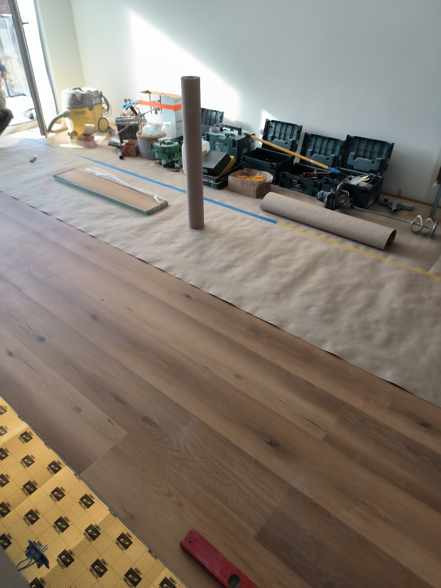 Montaż paneli podłogowych w jasnym wnętrzu, widoczne narzędzia, poziomica i podkład pod panele z nadrukiem 'CUTTING LINES', podłoga zabezpieczona papierem malarskim.