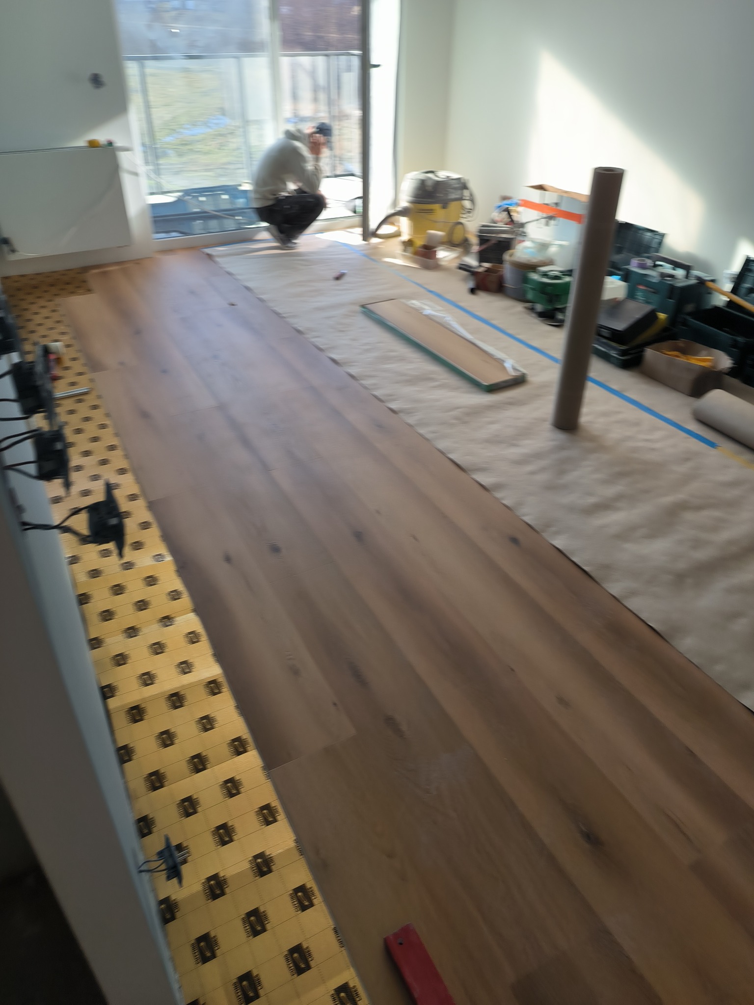 Układanie paneli podłogowych z widoczną warstwą izolacyjną; w tle pracownik w trakcie montażu, narzędzia i odkurzacz przemysłowy.