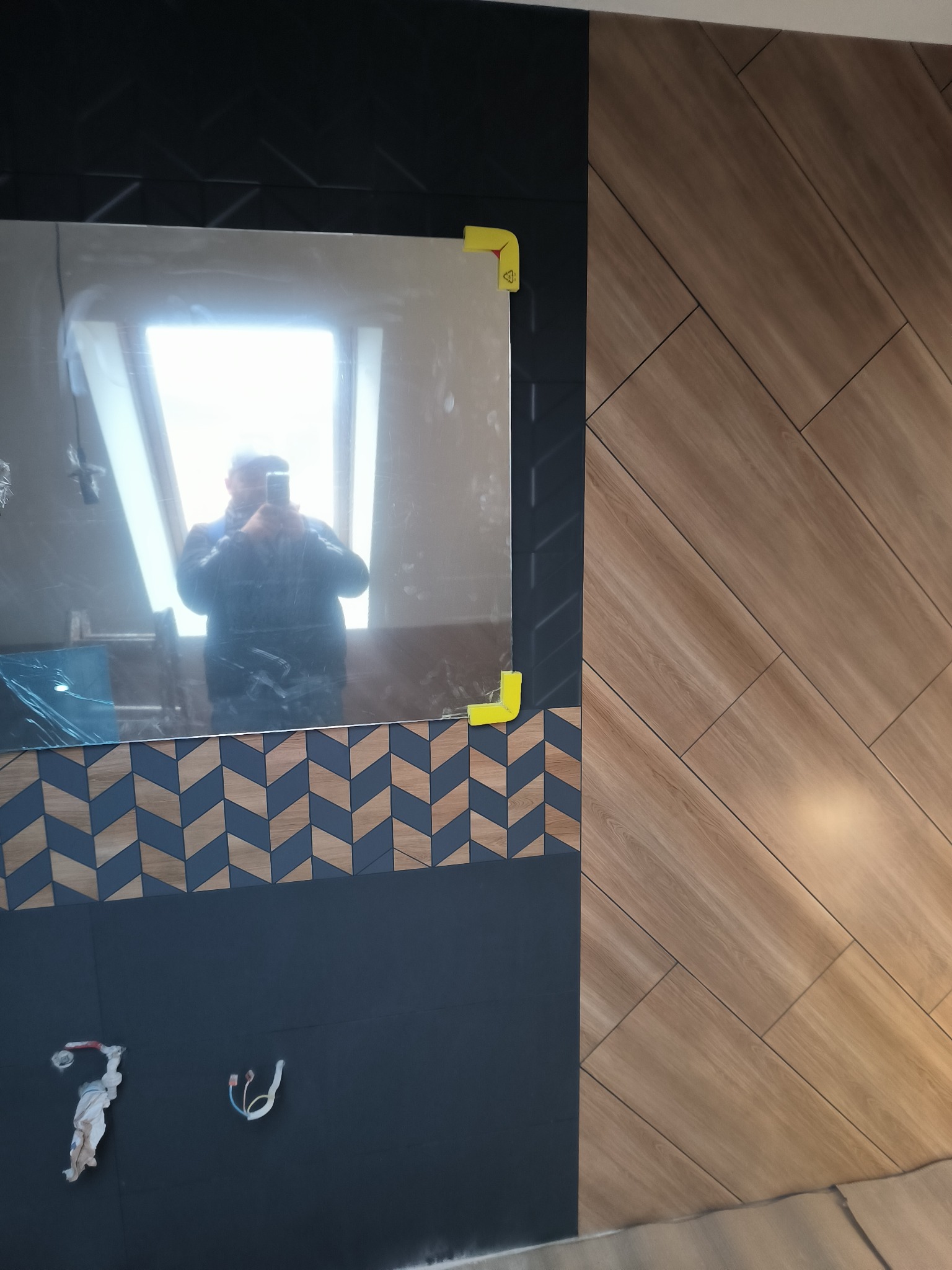 Fragment łazienki w trakcie wykańczania: lustro z zabezpieczeniami, drewnopodobne płytki ułożone w jodełkę, czarna ściana z wystającymi przewodami.