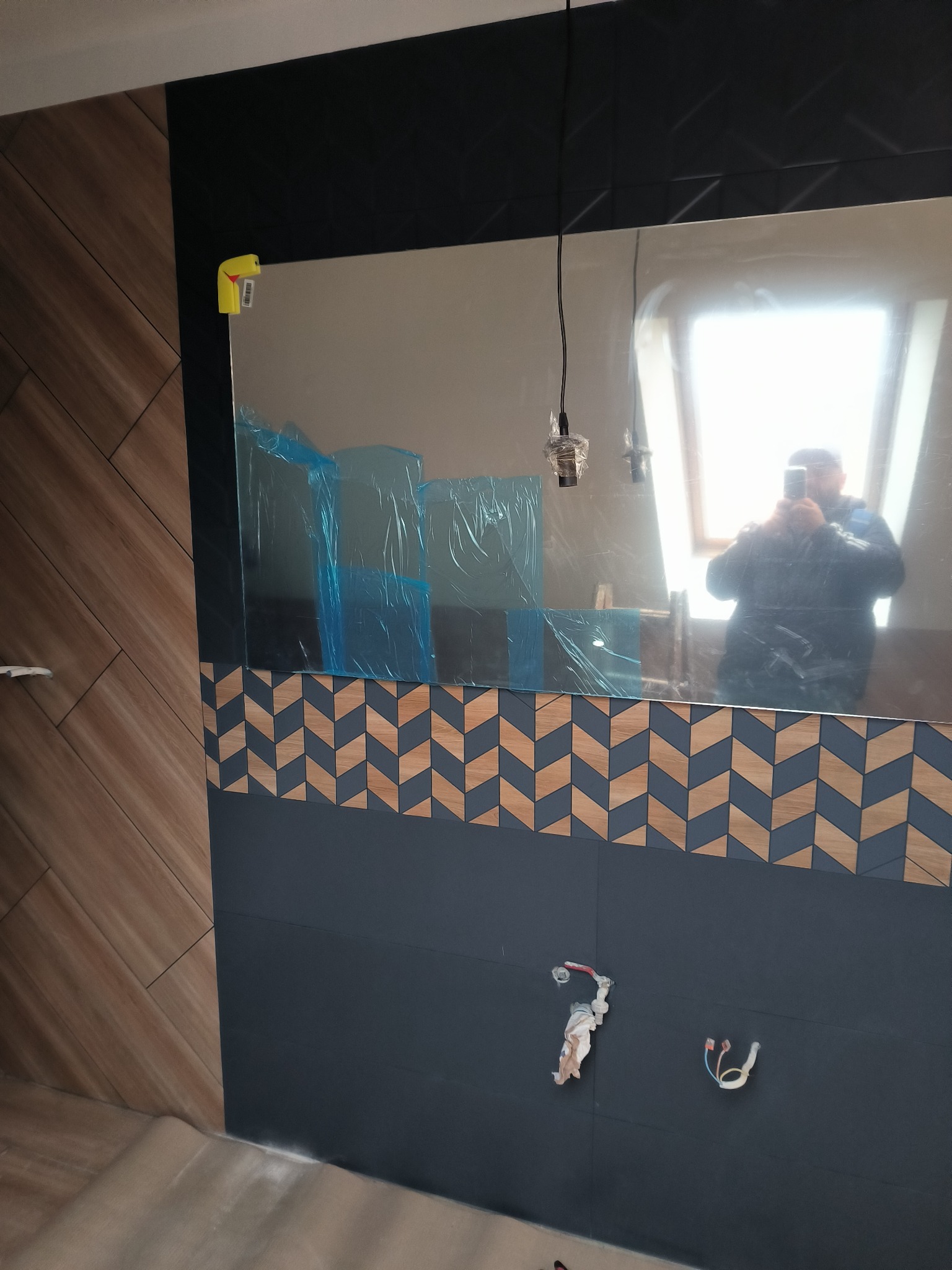 Fragment łazienki w trakcie remontu: ciemne płytki z drewnopodobnym wzorem chevron, lustro z folią ochronną i odbiciem fotografa, widoczne przewody elektryczne.