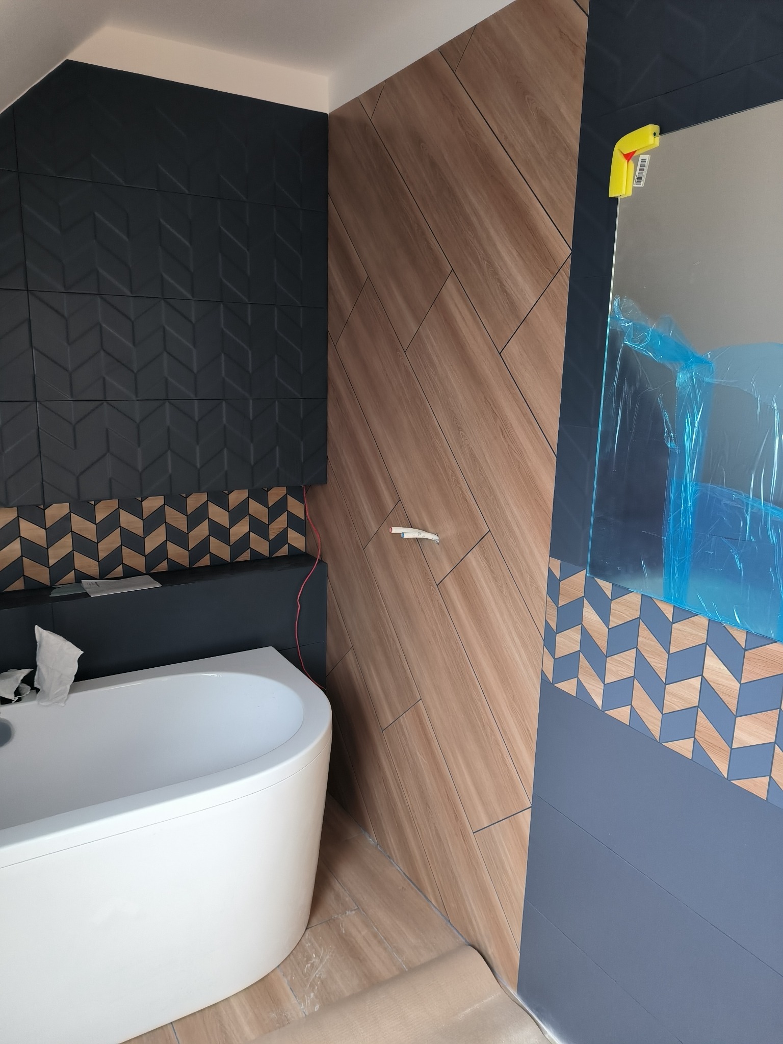 Nowoczesna łazienka w trakcie wykańczania z białą wanną narożną, ciemnymi płytkami z geometrycznym wzorem na ścianie i drewnopodobnymi płytkami ułożonymi pod skosem, lustro zabezpieczone folią.