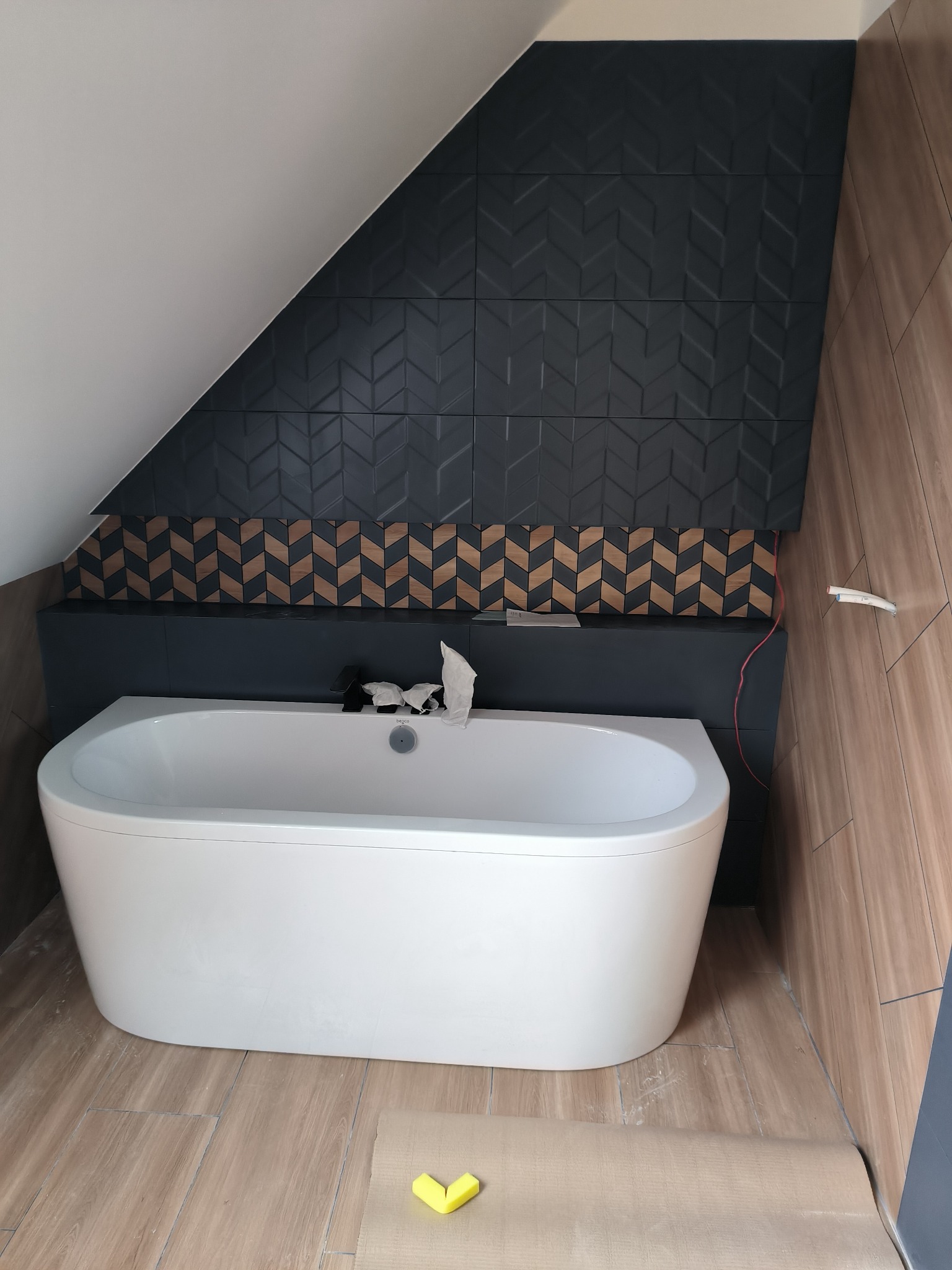 Biała wanna wolnostojąca na tle ściany z ciemnych, wzorzystych płytek w jodełkę i drewnopodobnych paneli, widoczne żółte zabezpieczenia na podłodze i wystające przewody.