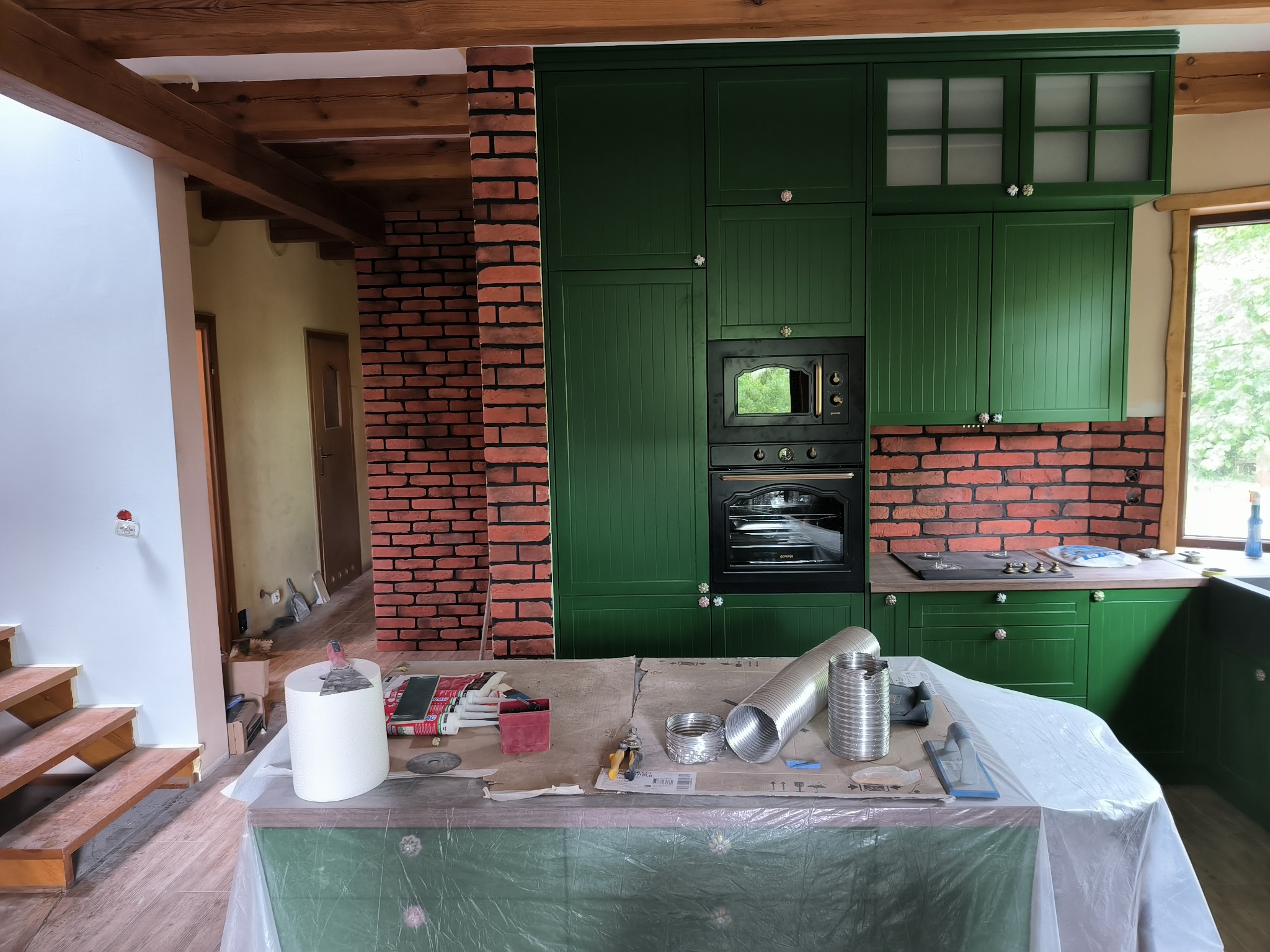 Wnętrze kuchni w trakcie prac wykończeniowych: zielone szafki, ceglana ściana, widoczne narzędzia i materiały na zabezpieczonej folią wyspie kuchennej, drewniane belki stropowe.
