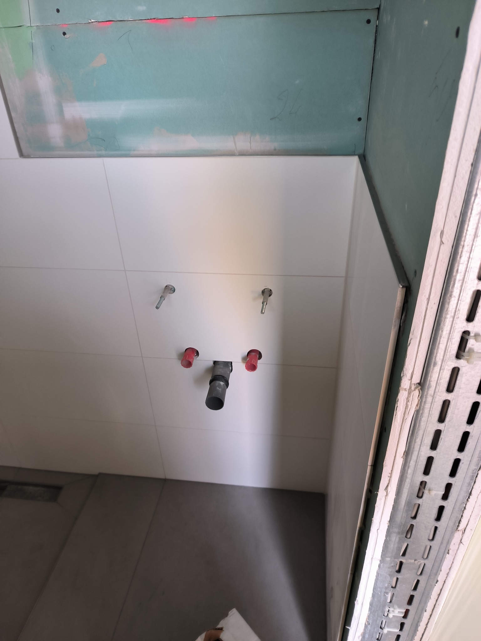 Świeżo położone, białe płytki na ścianie w łazience, widoczne wystające elementy instalacji wodnej, surowa ściana z płyt kartonowo-gipsowych w górnej części, metalowa konstrukcja nośna przy ścianie.