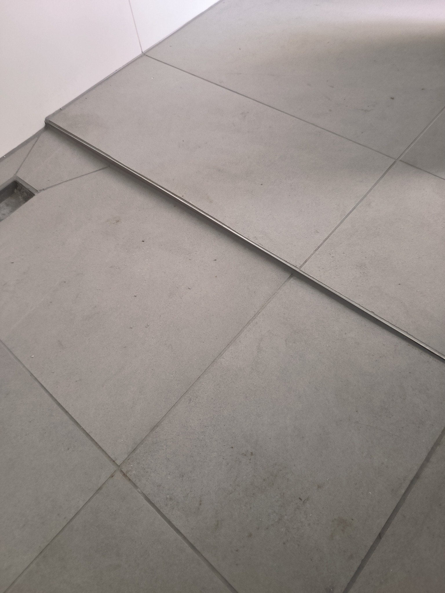 Szare płytki podłogowe z metalową listwą wykończeniową i odpływem liniowym, widoczne drobne zabrudzenia.