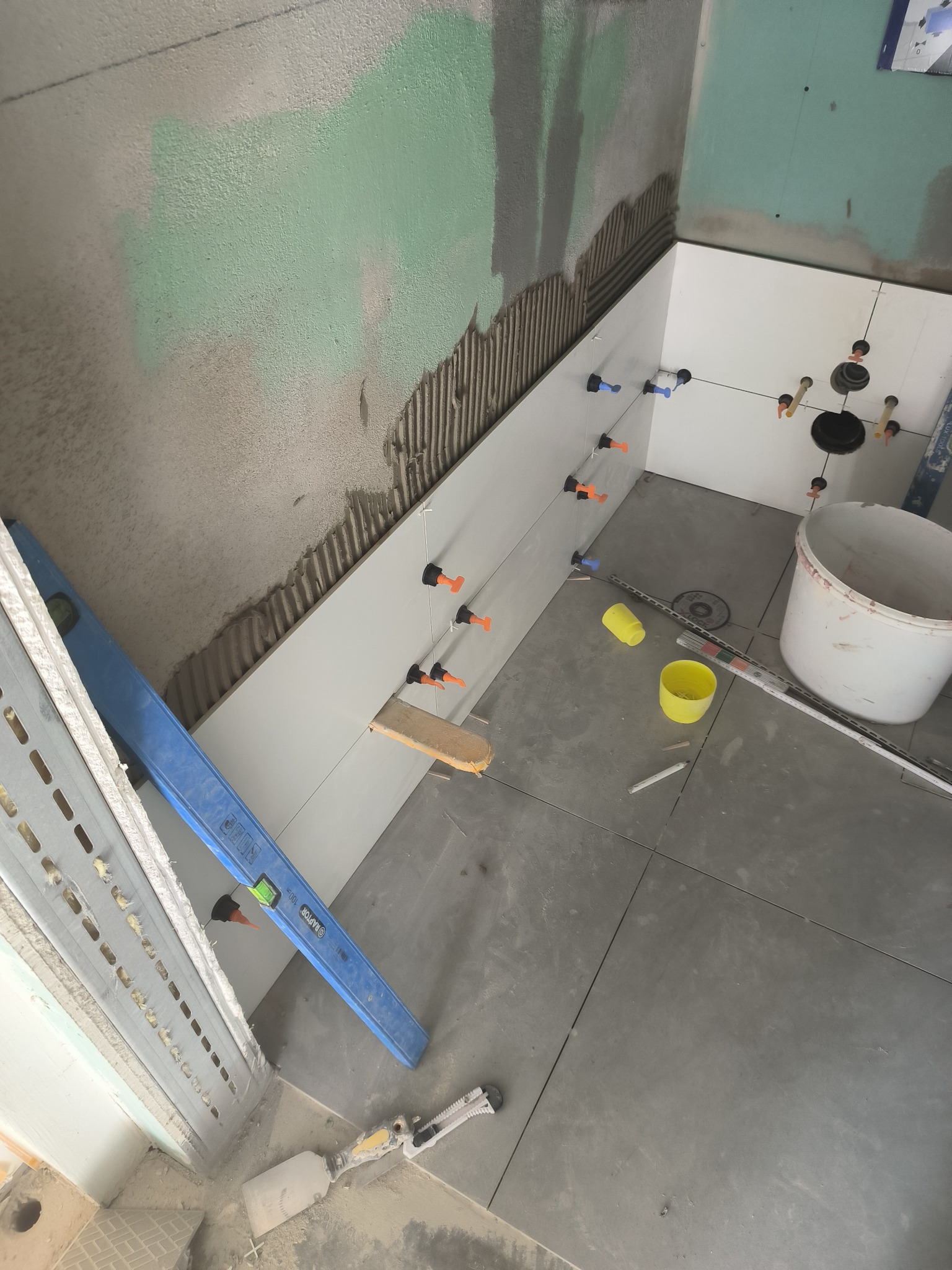 Białe płytki ścienne układane w łazience, widoczne poziomica, wiaderko, przyssawki do płytek i szpachelka, ściana częściowo przygotowana pod płytki.