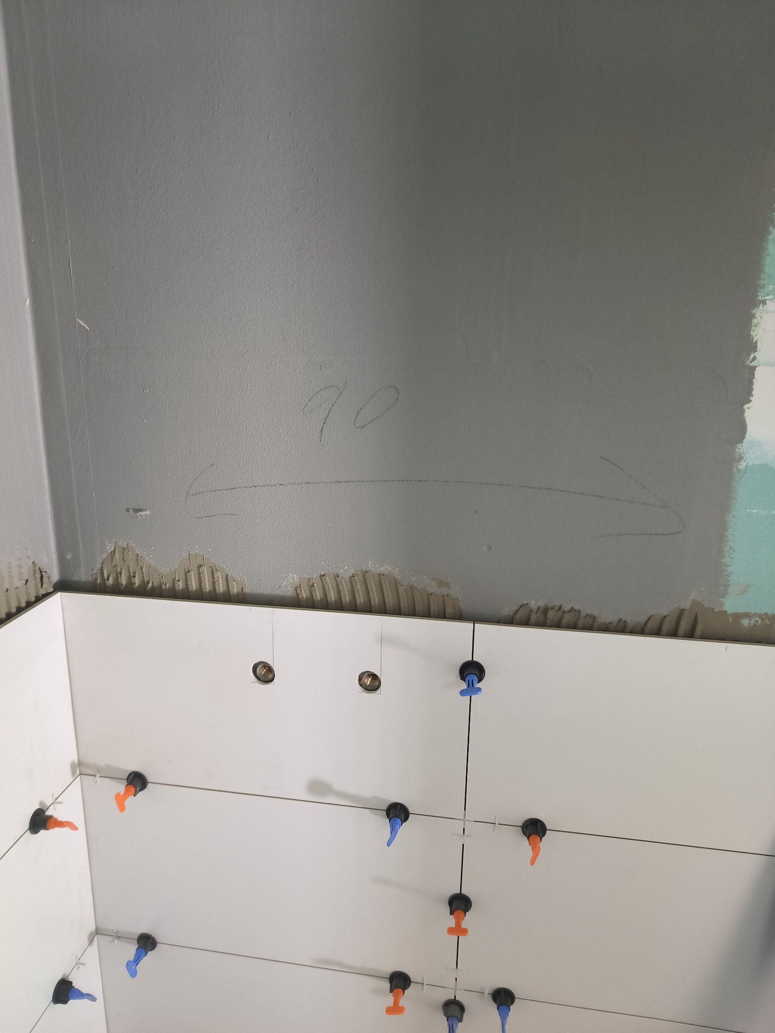 Białe płytki ścienne w trakcie układania, widoczne systemy poziomowania płytek (klipsy i kliny), ściana przygotowana pod płytki z zaznaczoną linią i wymiarem '70'.