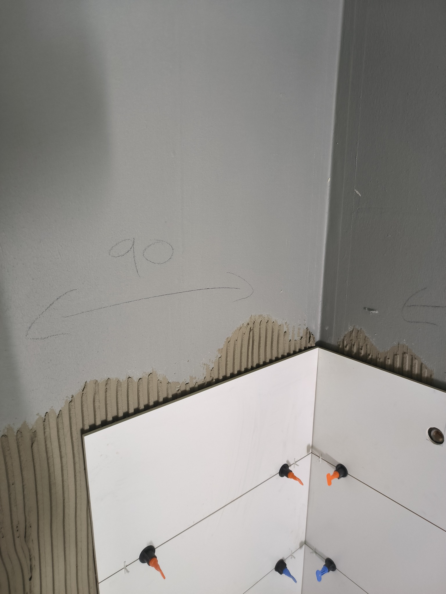 Układanie białych płytek w rogu pomieszczenia, widoczny klej na ścianie, system poziomowania płytek z kolorowymi klinami, notatka ołówkiem na ścianie.
