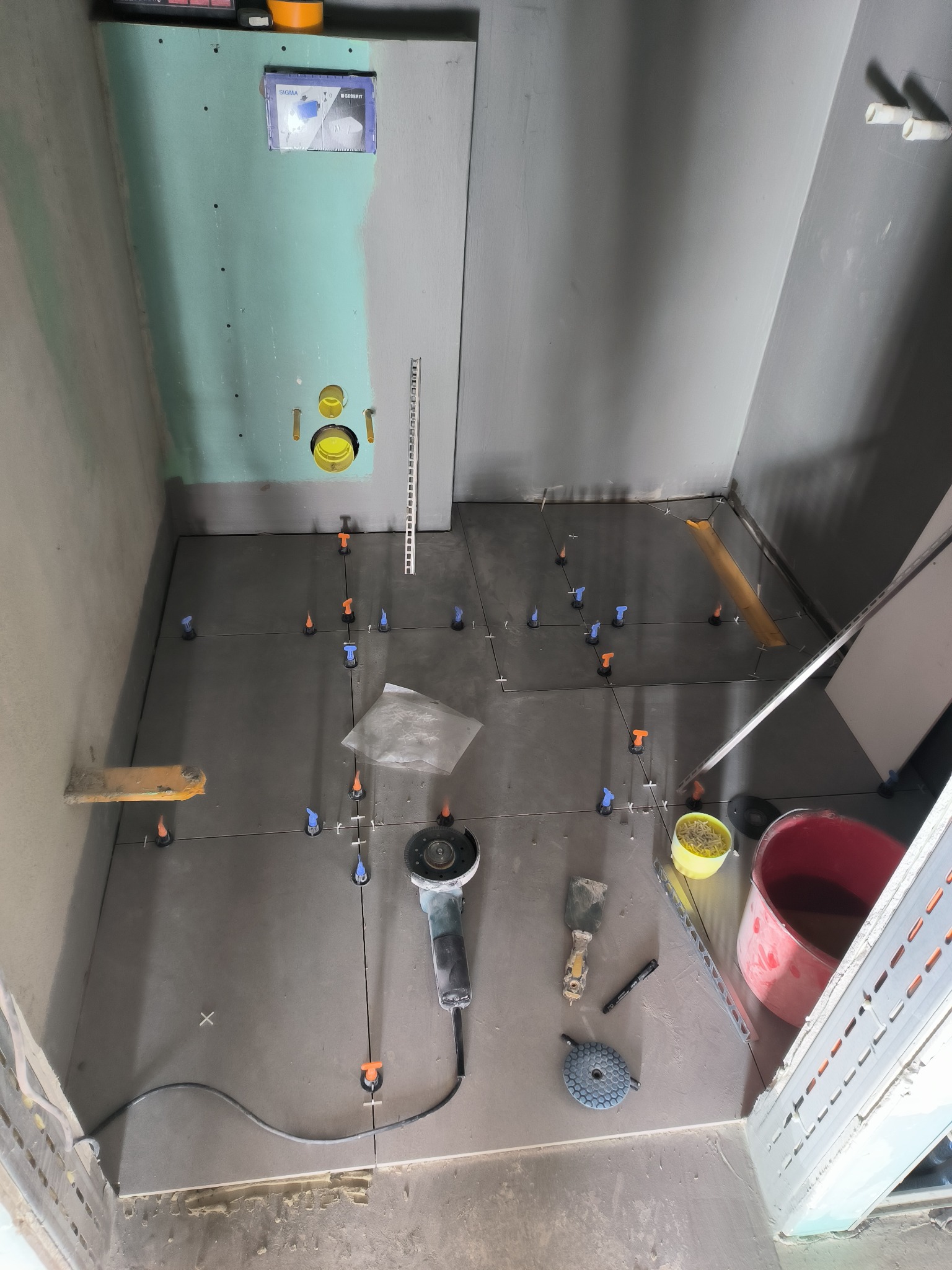 Układanie szarych płytek podłogowych w łazience, widoczne narzędzia: szlifierka kątowa, kielnia, poziomica, krzyżyki dystansowe i system poziomowania płytek.