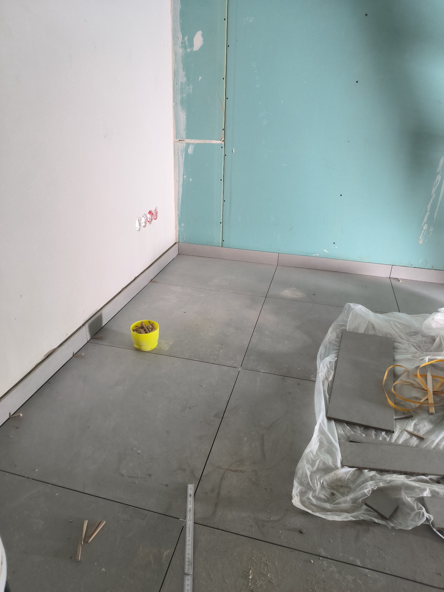 Układanie szarych płytek podłogowych w pomieszczeniu z częściowo wykończonymi ścianami, widoczne krzyżyki dystansowe, miarka i wiadro z klinami.