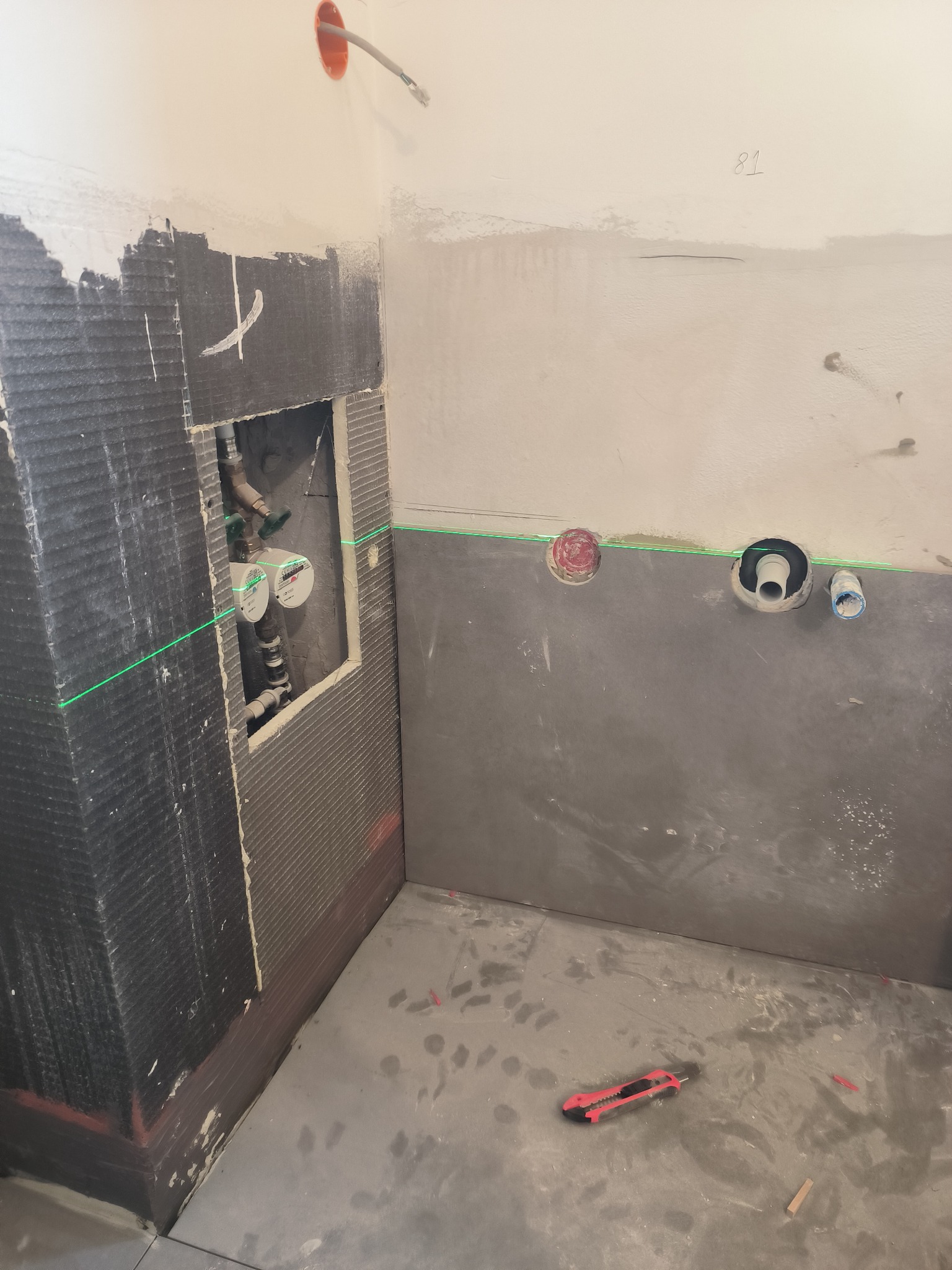 Przygotowanie ścian w łazience do położenia płytek, widoczne hydroizolacja, piony i poziomice laserowe, wystające rury i liczniki oraz nóż monterski na podłodze.