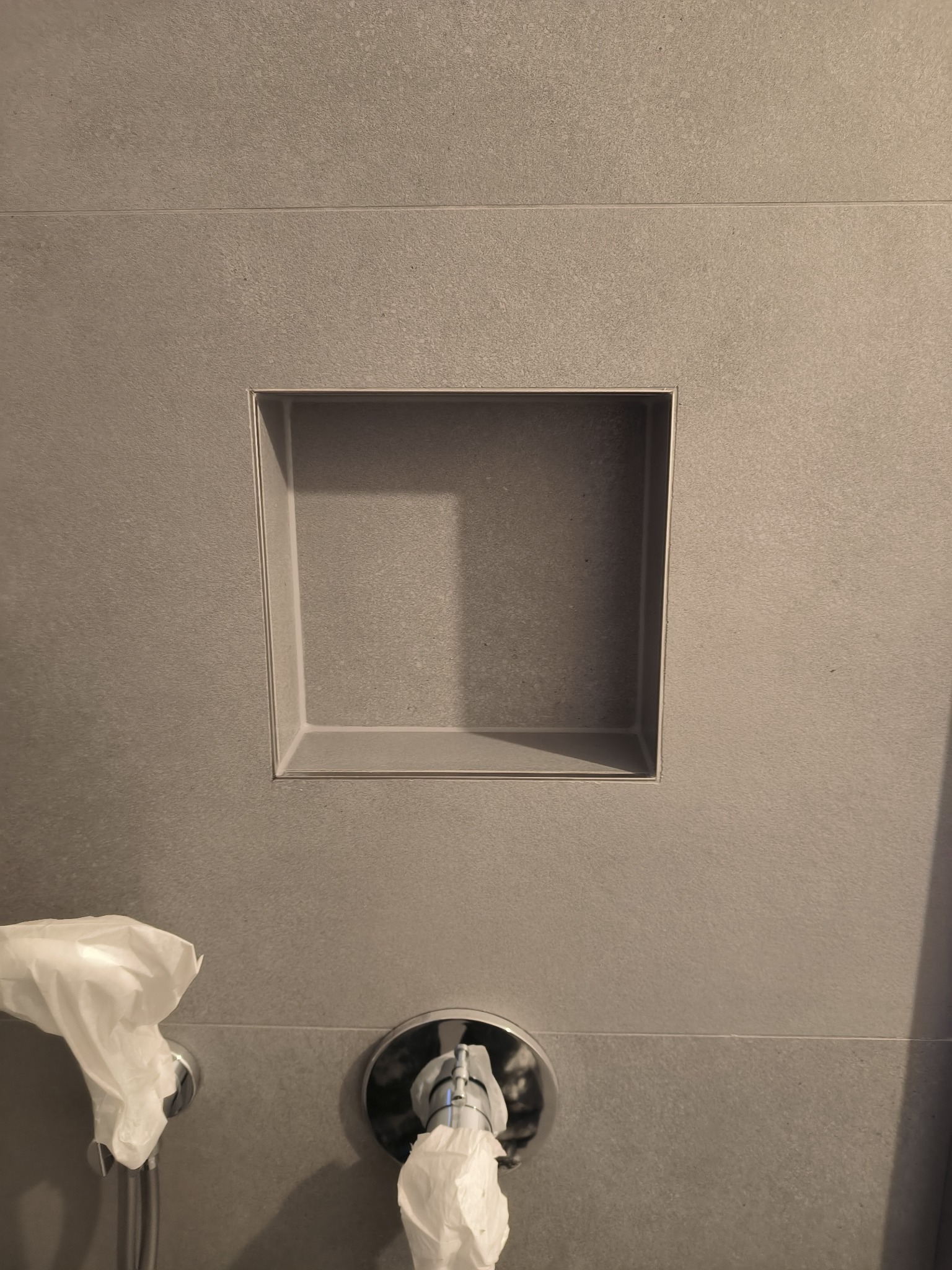 Szara ściana w łazience z wnęką wyłożoną płytkami, widoczna bateria z zawiniętą folią ochronną. Świeżo położone płytki.