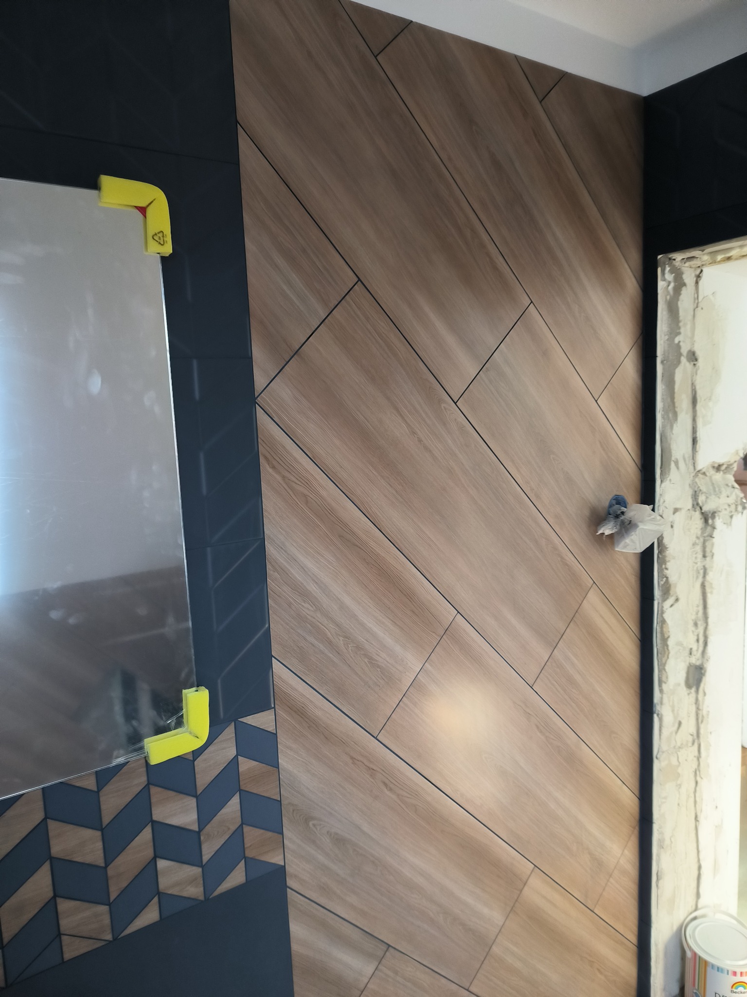 Ściana wyłożona płytkami imitującymi drewno w jodełkę, z fragmentem lustra zabezpieczonego żółtymi narożnikami i widocznym fragmentem surowego muru przy drzwiach.