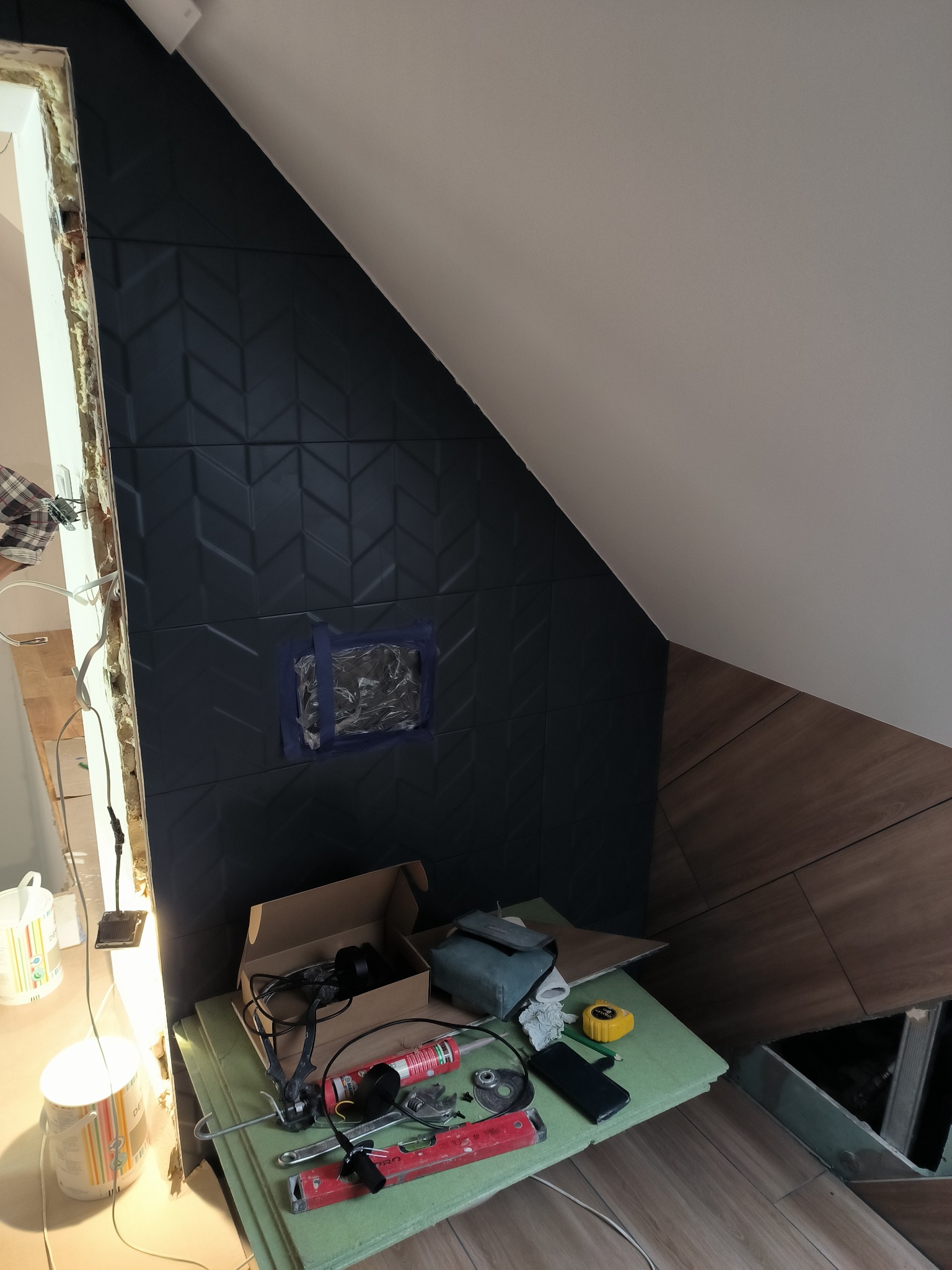 Montaż ciemnych, trójwymiarowych płytek na ścianie ze skośnym sufitem, zabezpieczony otwór taśmą, narzędzia i materiały robocze na prowizorycznym stole.