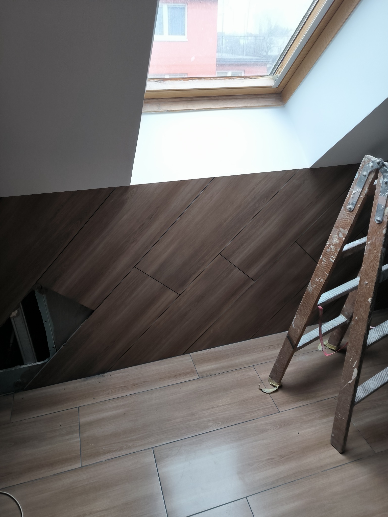 Ściana i podłoga wyłożone brązowymi płytkami imitującymi drewno, ułożonymi pod kątem, z widocznym otworem instalacyjnym i drewnianą drabiną opartą o ścianę, oświetlone światłem z okna dachowego.