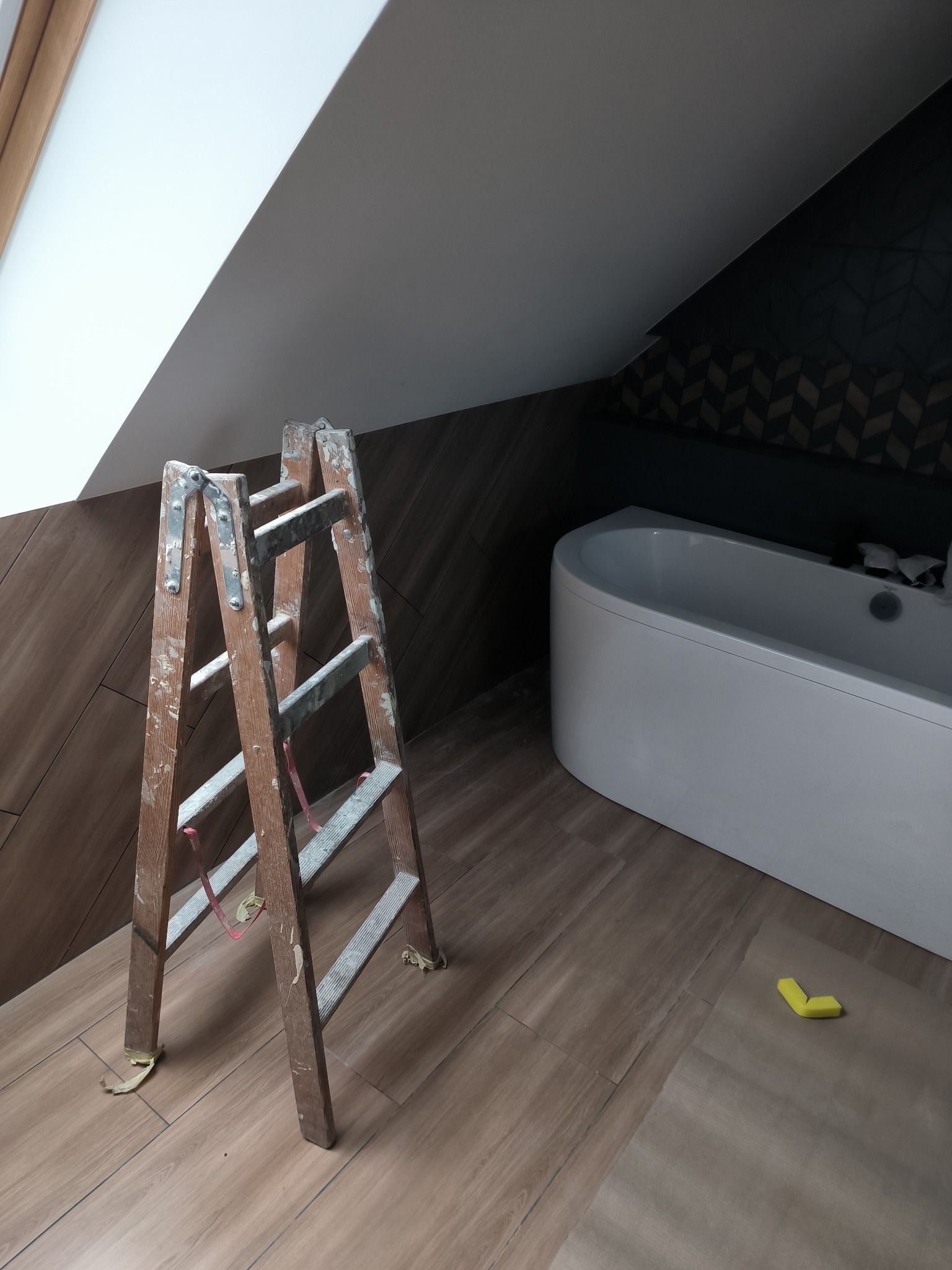 Poddasze łazienki w trakcie prac wykończeniowych: drewnopodobne płytki na podłodze i ścianach, biała wanna wolnostojąca, drabina malarska z resztkami farby i zabezpieczona folia na podłodze.