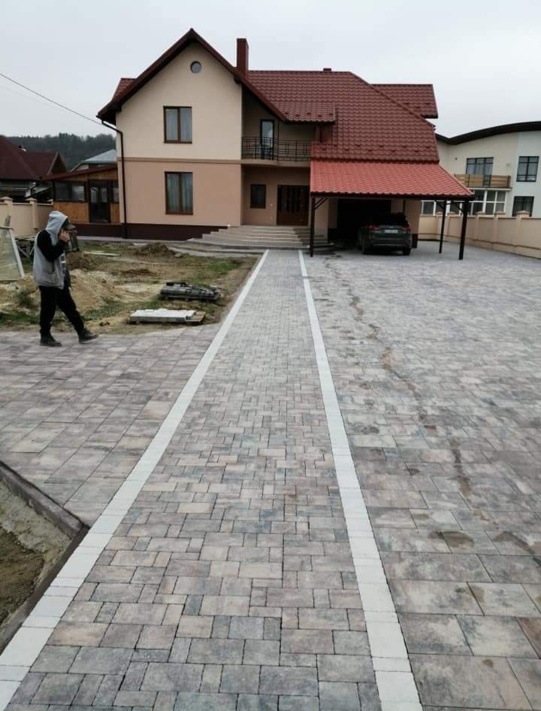 Ukończony podjazd z kostki brukowej w odcieniach szarości z białymi pasami, prowadzący do domu z czerwonym dachem i wiatą garażową, osoba w bluzie z kapturem przechadza się obok.