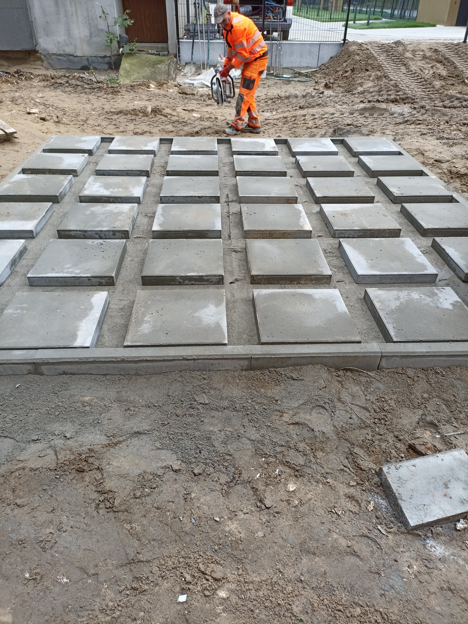 Przygotowane podłoże z betonowych płyt i obrzeży, pracownik w pomarańczowym kombinezonie przycina płytę piłą spalinową w tle.