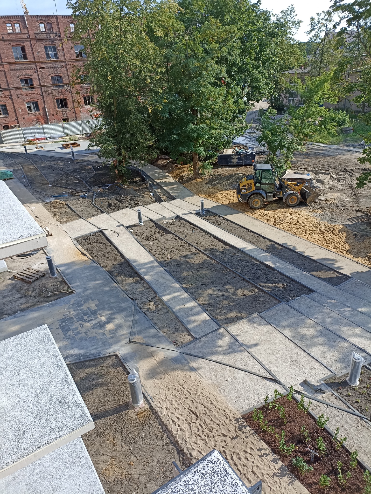 Widok z góry na teren budowy z przygotowanymi korytarzami pod nawierzchnię, gdzie w tle widoczna jest koparko-ładowarka i budynek z czerwonej cegły.