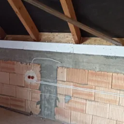 Surowy stan poddasza w trakcie prac: widoczne ceglane ściany z bruzdami, wystające przewody elektryczne z puszkami, drewniana konstrukcja dachu z izolacją.