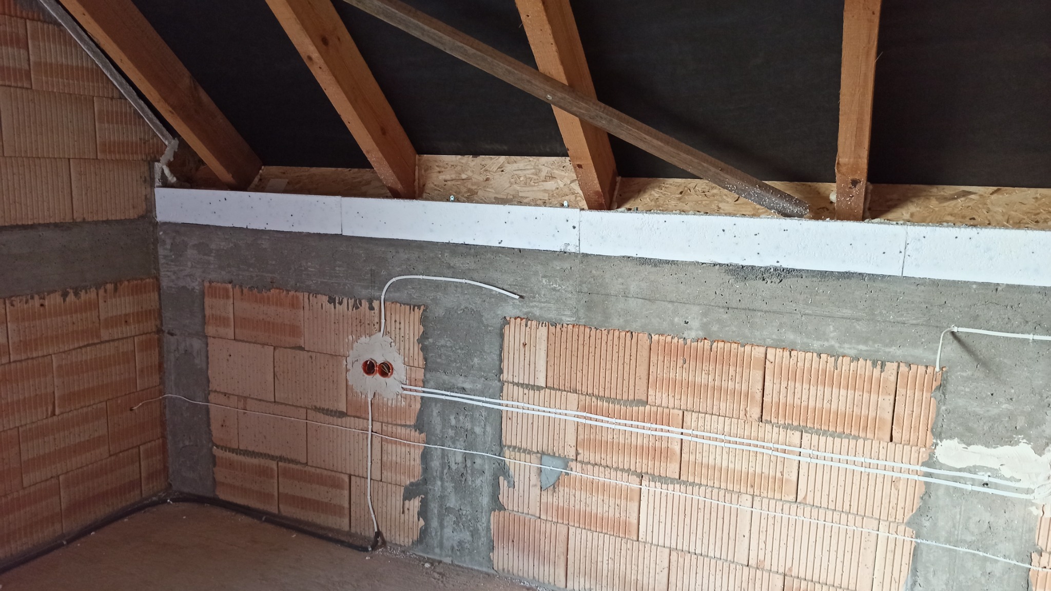 Surowy stan poddasza w trakcie prac: widoczne ceglane ściany z bruzdami, wystające przewody elektryczne z puszkami, drewniana konstrukcja dachu z izolacją.