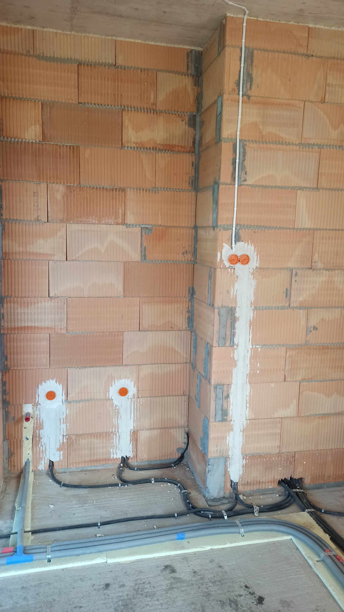 Surowy mur z pustaków ceramicznych z wyznaczonymi punktami pod gniazdka elektryczne, bruzdami wypełnionymi gipsem, peszlami z przewodami i rurami instalacyjnymi na podłodze.