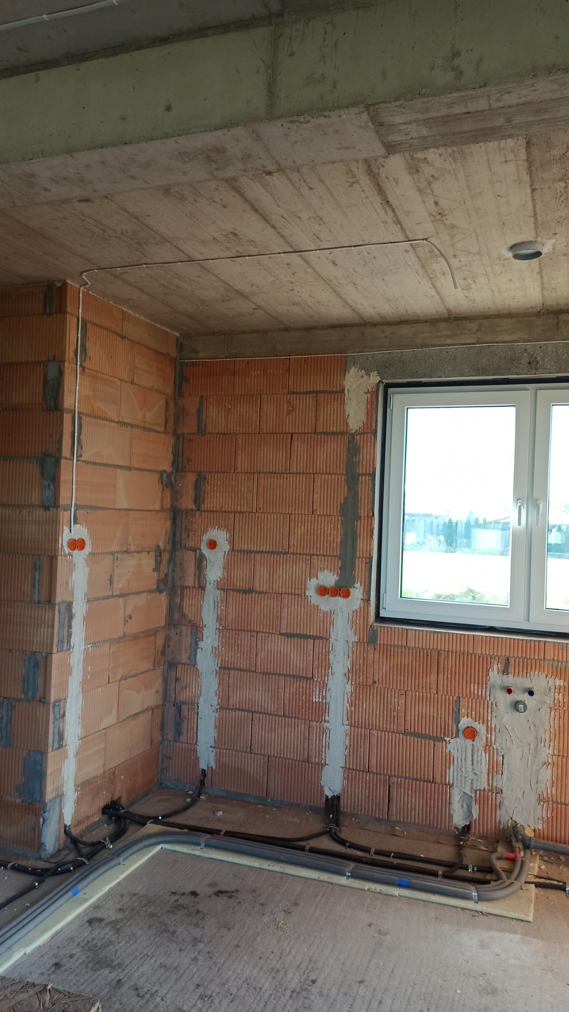 Wnętrze w stanie surowym: instalacja elektryczna w trakcie montażu, widoczne bruzdy w ceglanej ścianie z zamontowanymi puszkami elektrycznymi i przewodami w peszlach, pod stropem poprowadzony kabel.
