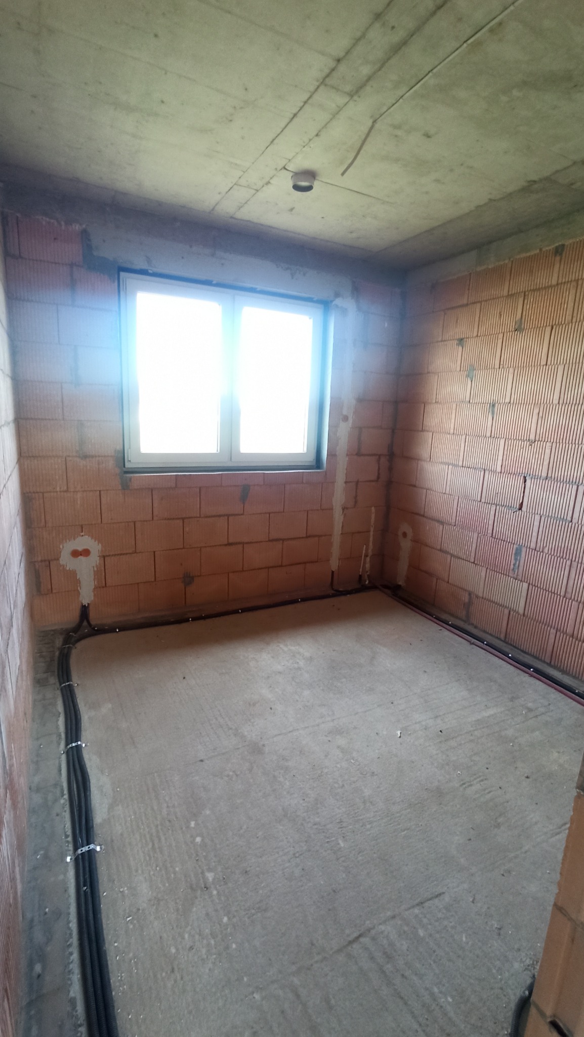 Wnętrze surowego domu jednorodzinnego w trakcie instalacji elektrycznej: widoczne przewody w peszlach mocowane do ściany z czerwonej cegły, puszki instalacyjne, surowy betonowy sufit z punktem...