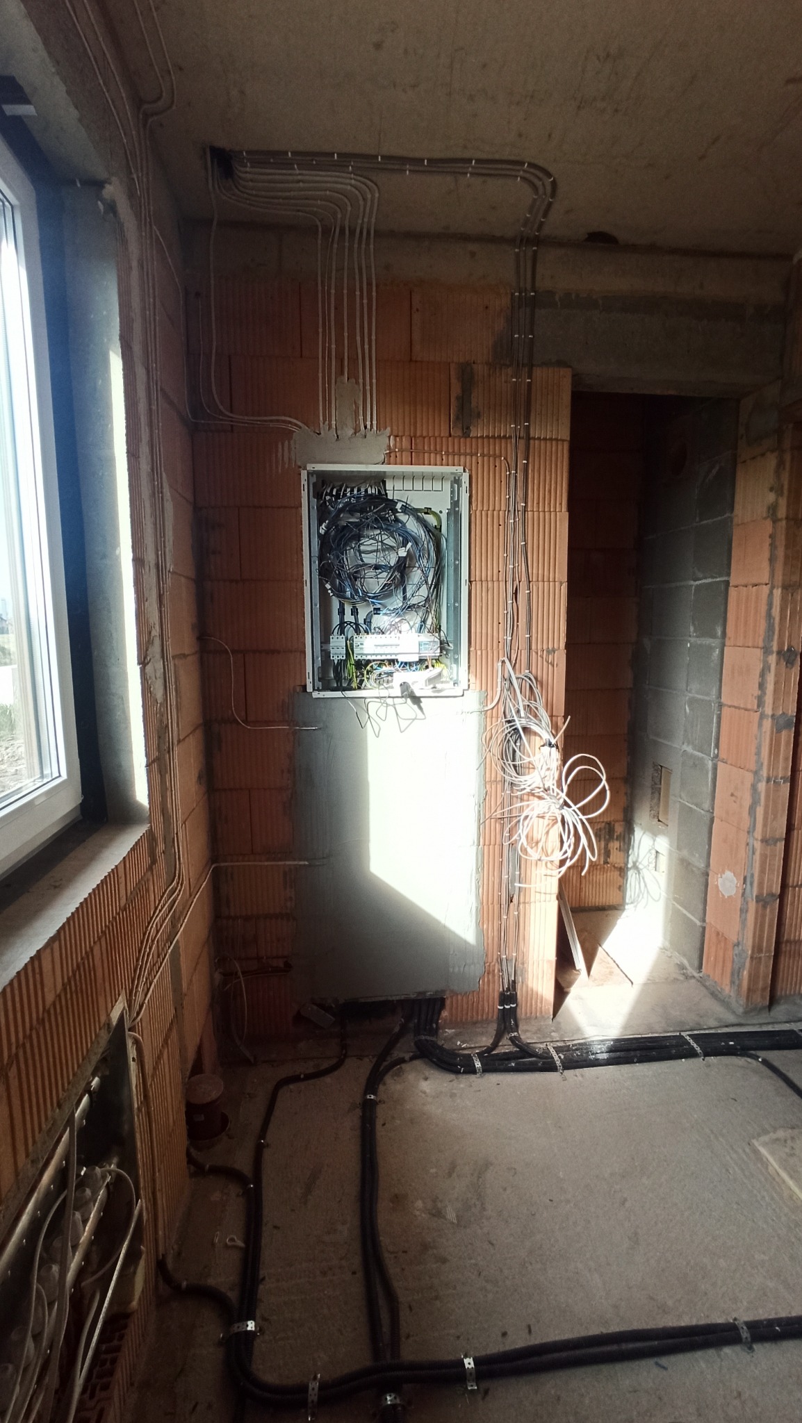 Wnętrze w stanie surowym z widoczną rozdzielnicą elektryczną w trakcie montażu, liczne przewody rozprowadzone po ścianach i podłodze, częściowo zamaskowane szarym tynkiem.
