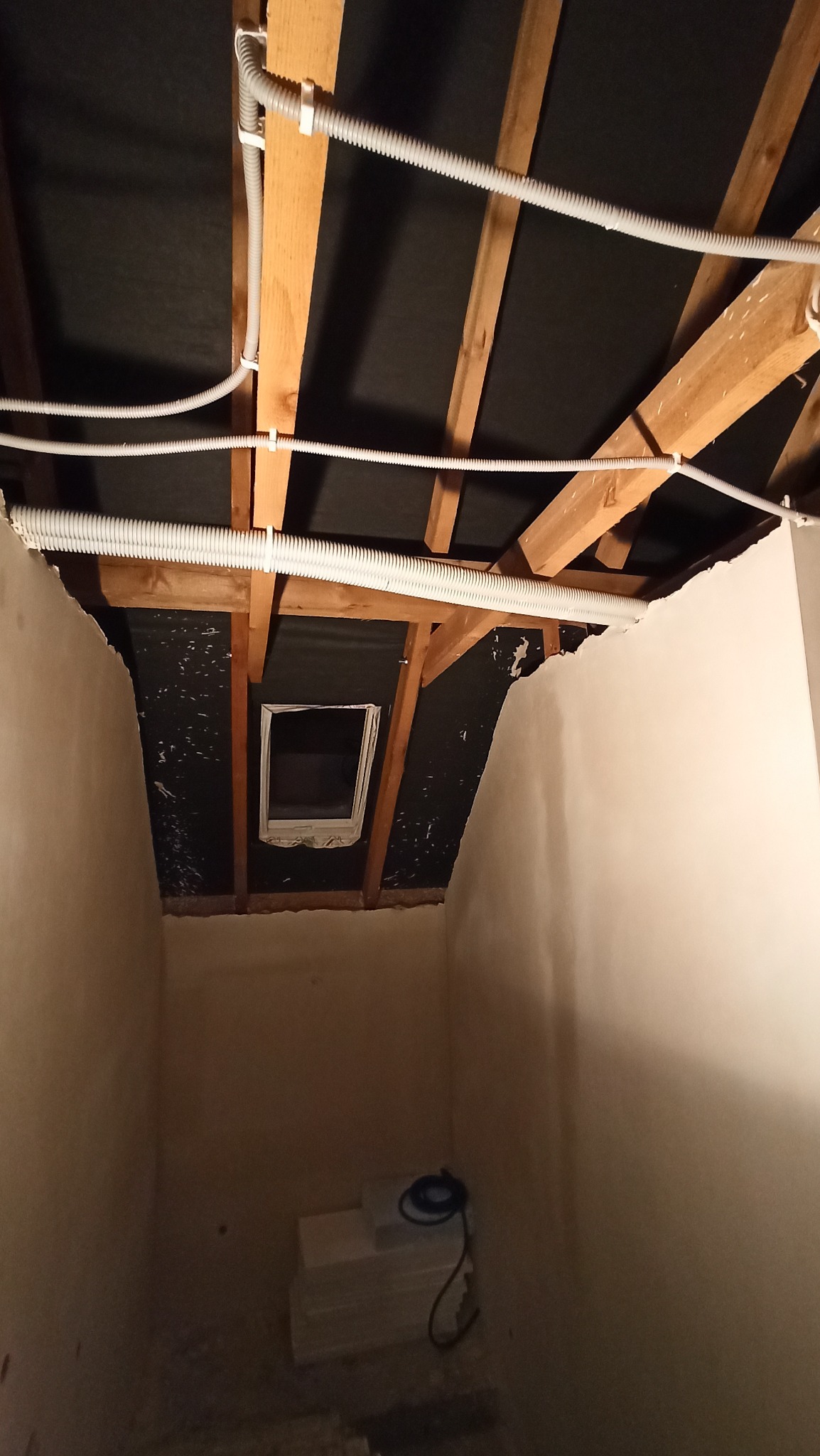 Poddasze w trakcie prac wykończeniowych, widoczne przewody elektryczne w peszlach mocowane do drewnianej konstrukcji dachu, ściany w stanie surowym, okno dachowe.