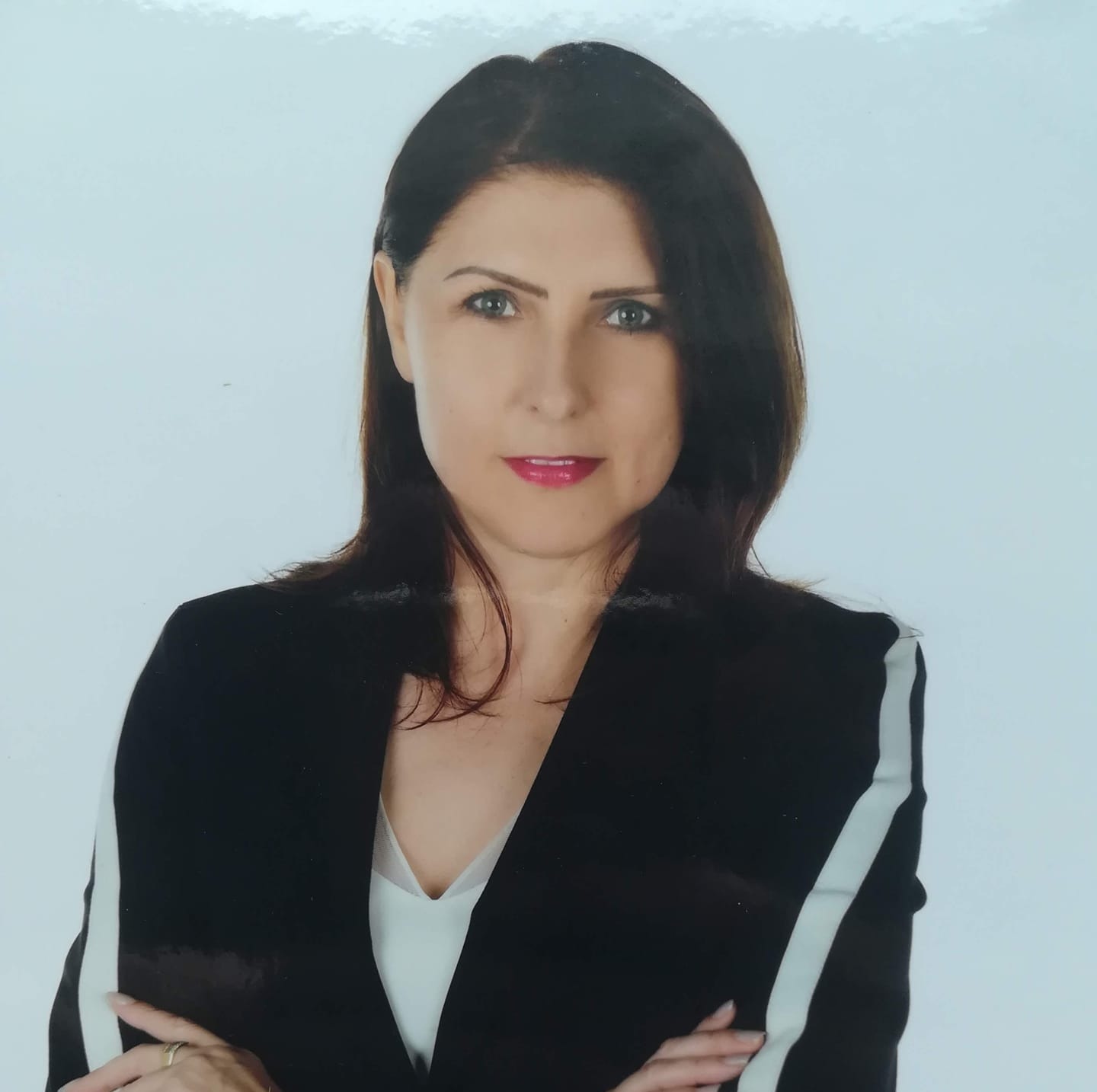 Portret kobiety w czarnym żakiecie z białym paskiem, skrzyżowane ręce, pierścionek na palcu, na jasnym tle.