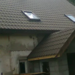 Fragment dachu z dachówki trapezowej w odcieniach szarości, z dwoma oknami dachowymi, widok na komin i częściowo otynkowaną ścianę budynku w trakcie budowy, z widocznymi rusztowaniami.