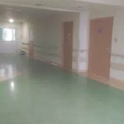 Długi, słabo oświetlony korytarz z zieloną podłogą i rzędem zamkniętych, drewnianych drzwi z tabliczkami informacyjnymi. Na końcu korytarza widoczne przeszklone drzwi.