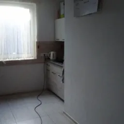 Kuchnia z oknem i firanką, widoczny fragment lodówki, białe szafki, czajnik elektryczny i kalendarz na ścianie.