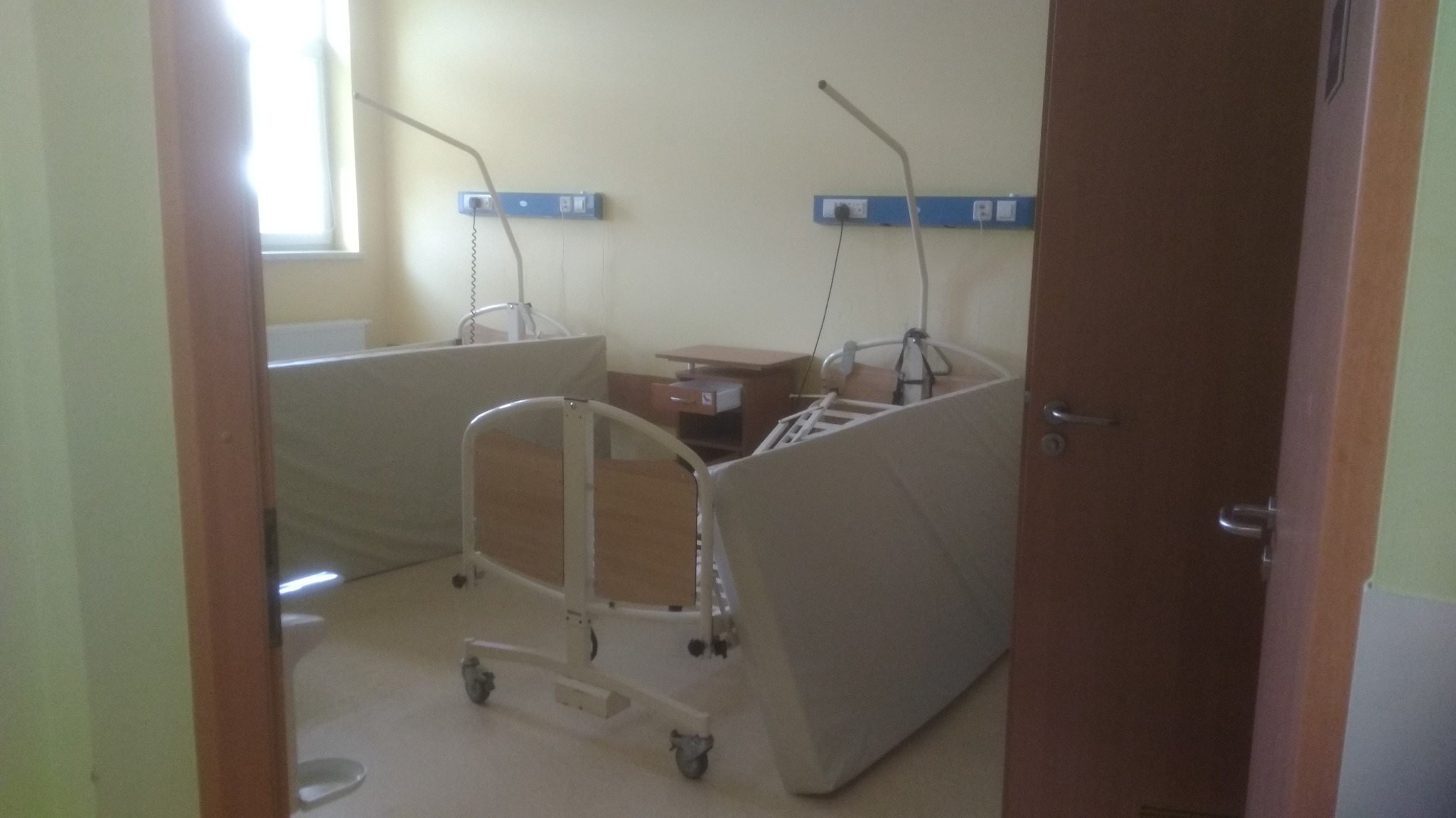 Widok przez otwarte drzwi na salę szpitalną z dwoma łóżkami z materacami, szafką nocną i panelami ściennymi z gniazdkami, oświetloną naturalnym światłem z okna.