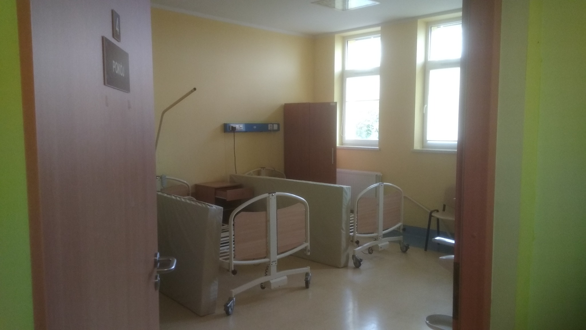 Pusty, jasny pokój szpitalny z dwoma łóżkami na kółkach, szafką nocną i szafą, widok zza uchylonych drzwi z napisem 'POKÓJ'.